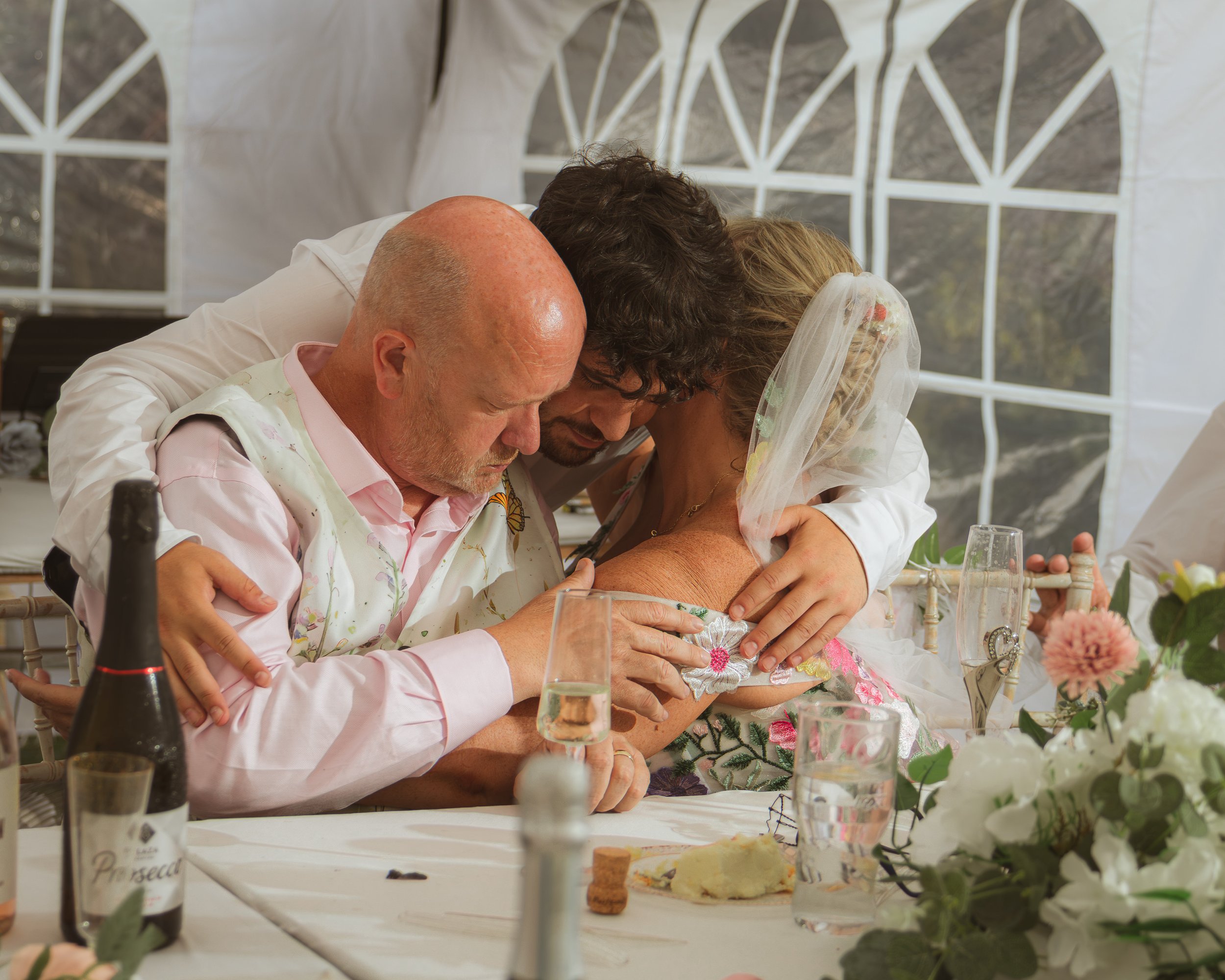 Three people hugging at a celebration table inside a decorated tent, with flowers, wine, and cake.
