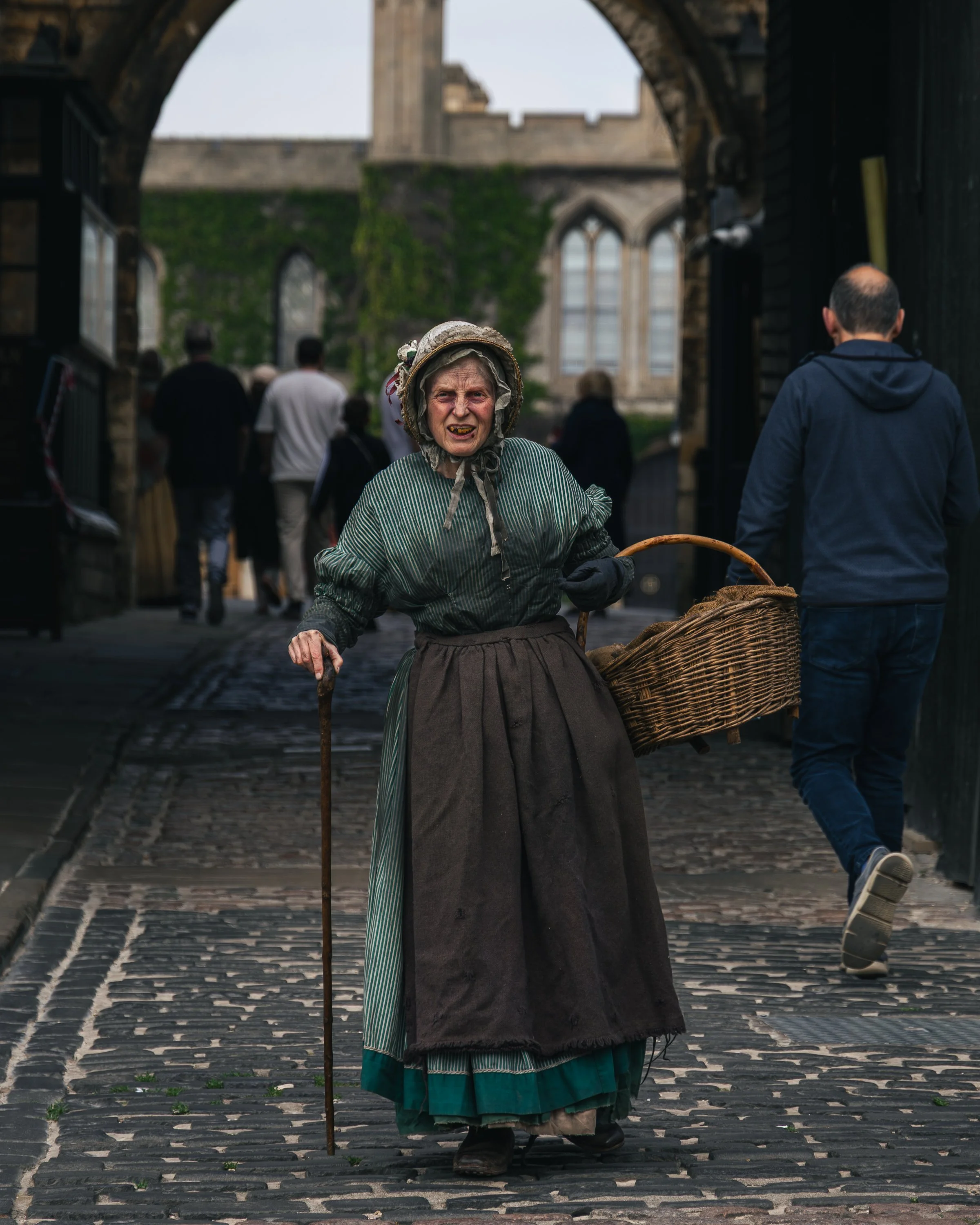 An elderly woman dressed in historical costume, holding a walking stick and carrying a wicker basket, walking on a cobblestone street under an archway with a stone building in background.