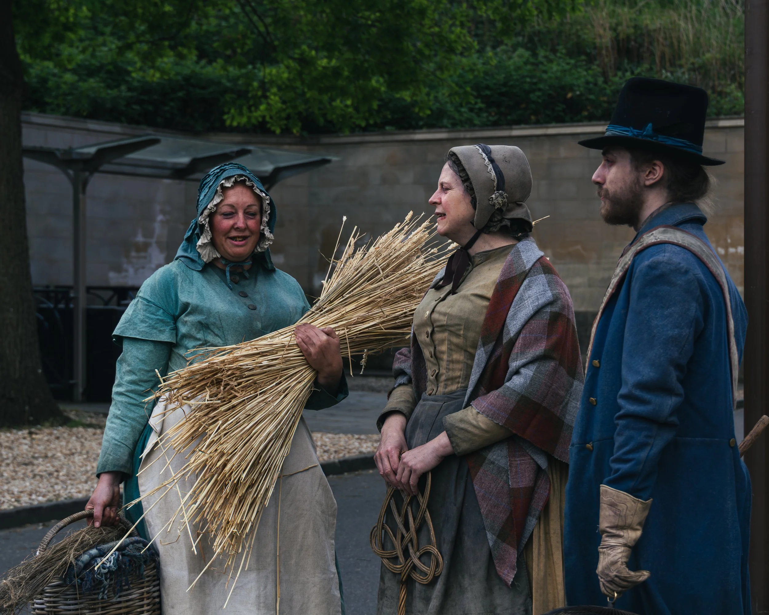 Three women dressed in historical clothing stand near each other outdoors, engaged in a conversation, with green trees and a stone wall in the background. One woman holds a bundle of straw, and a basket is also visible.