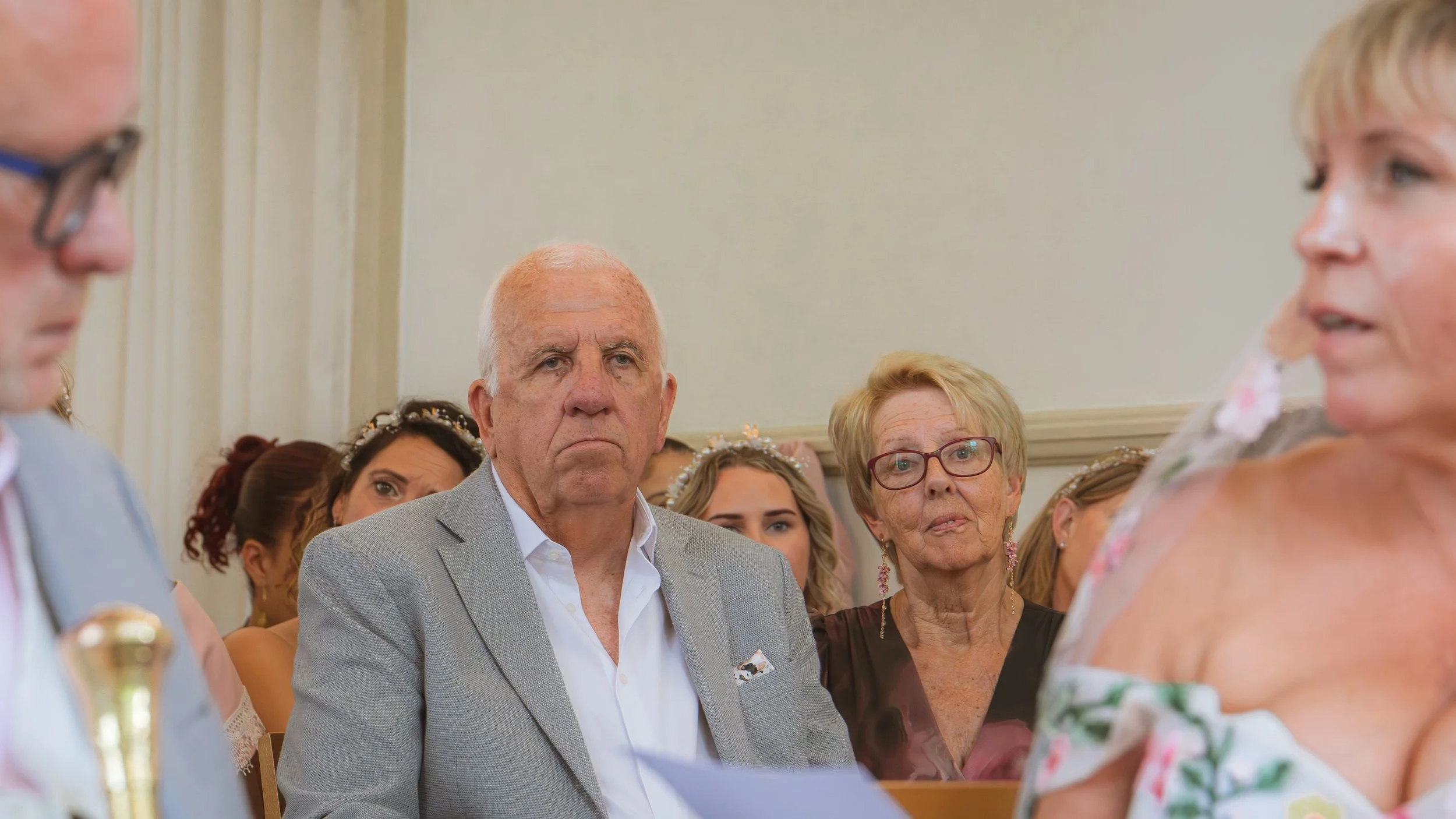 A group of people attending a formal event, with an elderly man in a light gray suit and a serious expression among them. Others in the background include an elderly woman with glasses and children, some wearing floral dresses and accessories.
