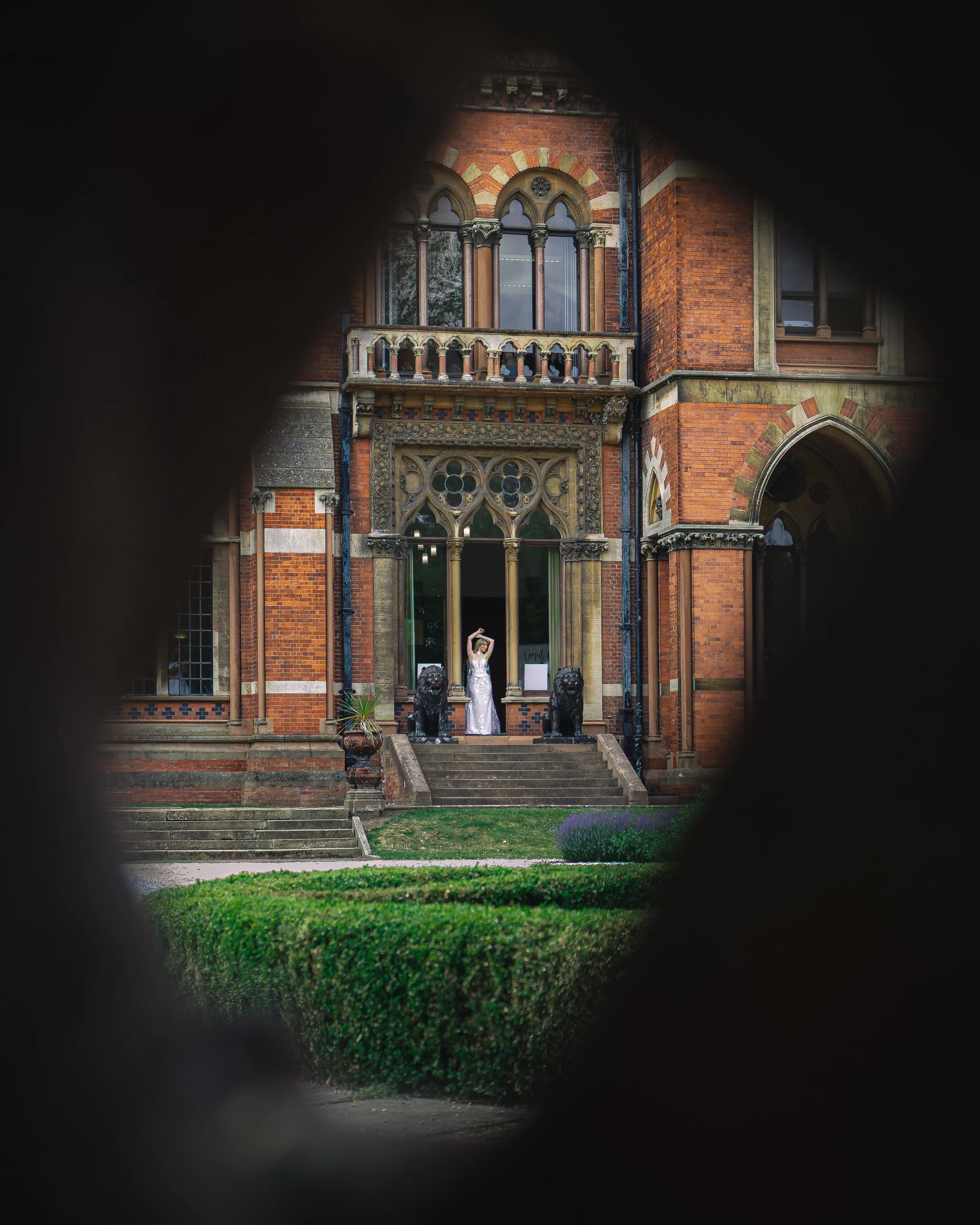 View of a historic brick building with Gothic architecture, including arched windows and detailed stonework. A woman in a wedding dress stands on the steps posing, flanked by two lion statues. The photo is taken through a small opening, framing the s