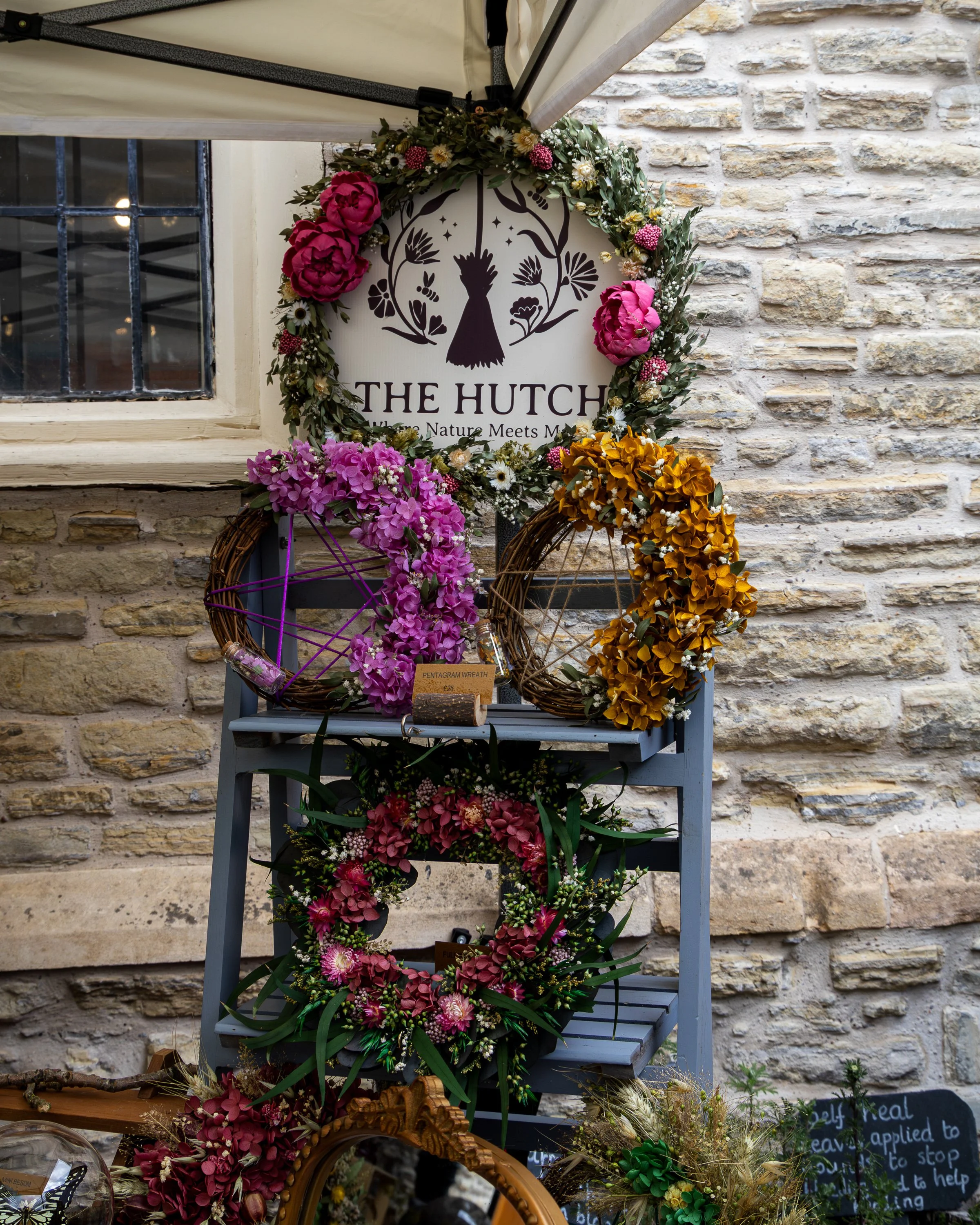 Decorative floral wreaths and arrangements on display in front of a stone wall and a sign that reads "The Hutch" with a bee logo.