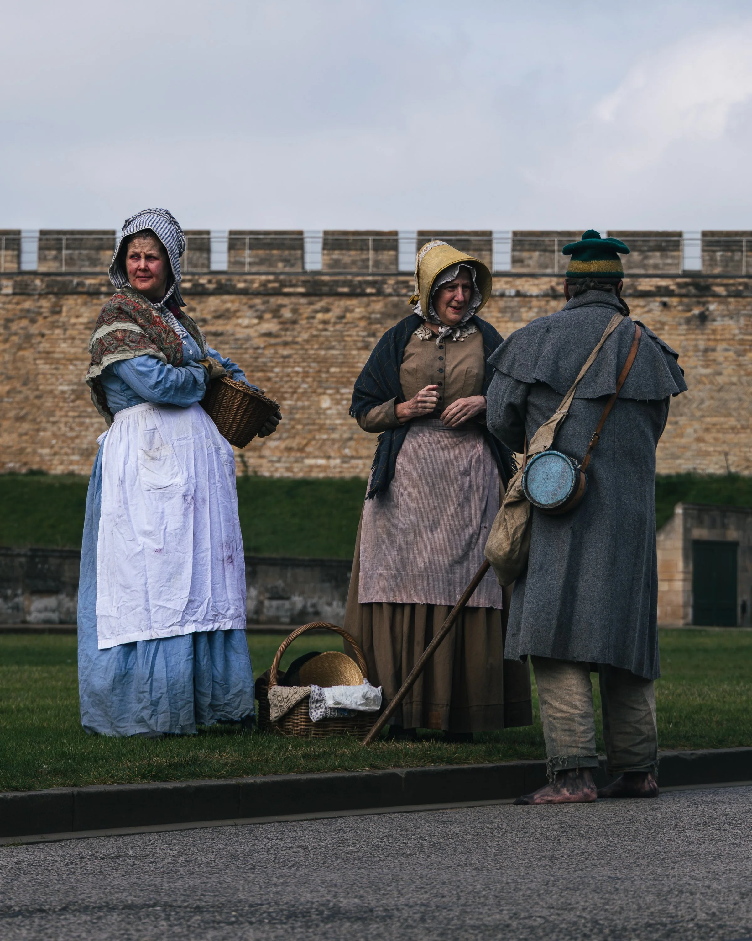 Three women dressed in historical clothing standing near a brick wall, with a basket and a hat at their feet, in an outdoor setting.