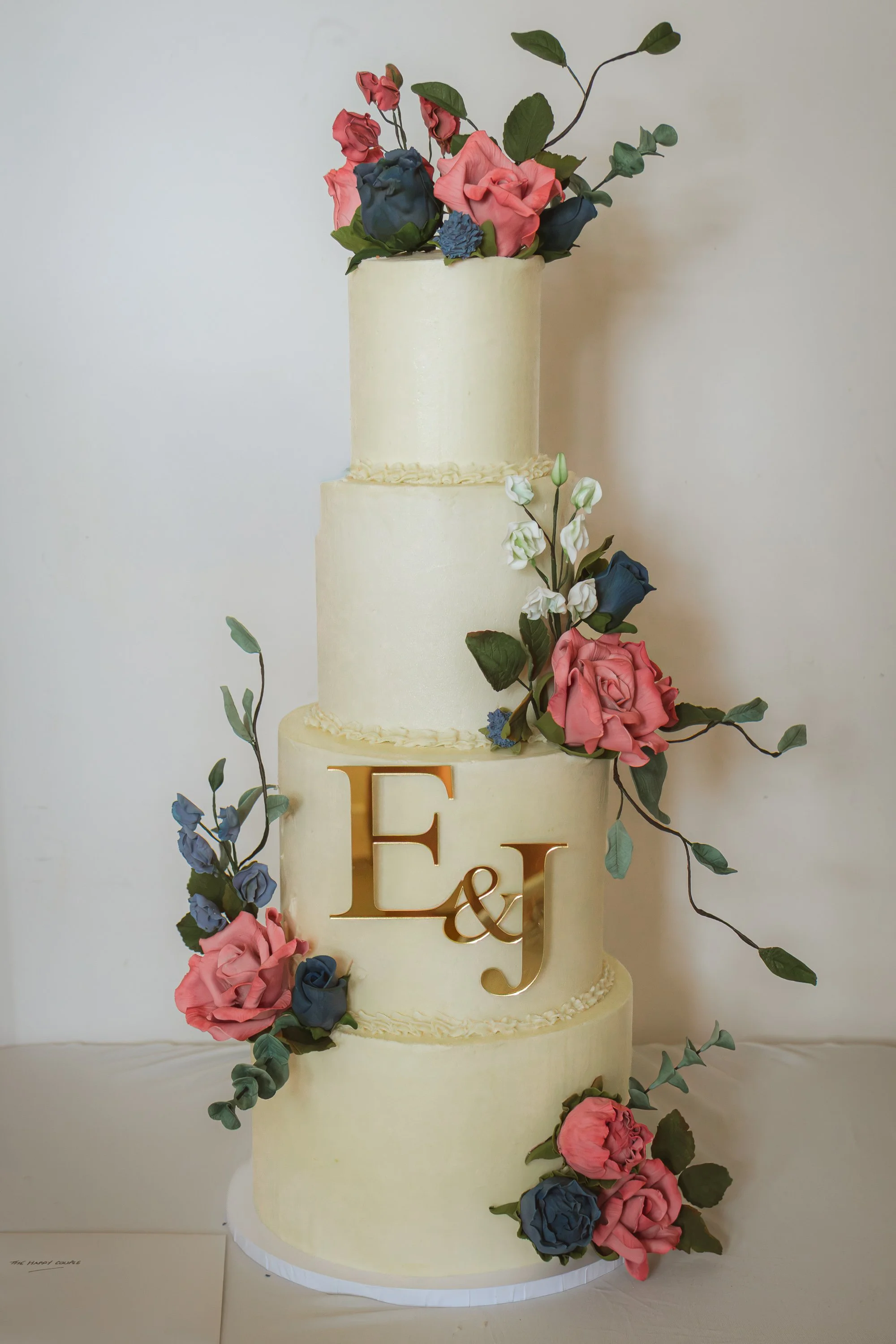 Three-tiered cream-colored wedding cake decorated with pink and blue sugar flowers and green leaves, with gold initials 'E & J' on the middle tier.