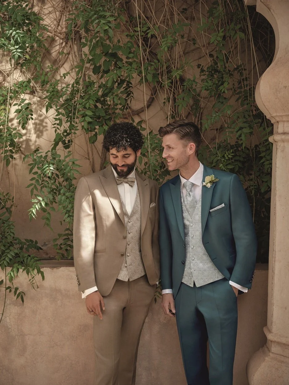 Zwei Männer in Hochzeitsanzügen stehen unter einer Bögen mit Kletterpflanzen und sprechen sich an, einer lächelt und der andere schaut verlegen.