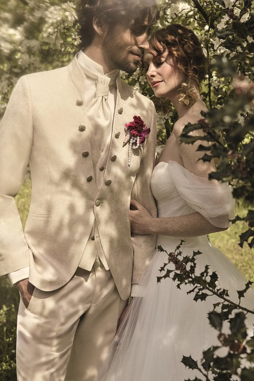 Ein lächelndes Paar in Hochzeitskleidung umarmt sich in einem Garten mit blühenden weißen Blumen. Der Mann trägt einen cremefarbenen Anzug mit silbernen Knöpfen, die Frau ein weißes Brautkleid mit Tüll und große Blumenohrringe.