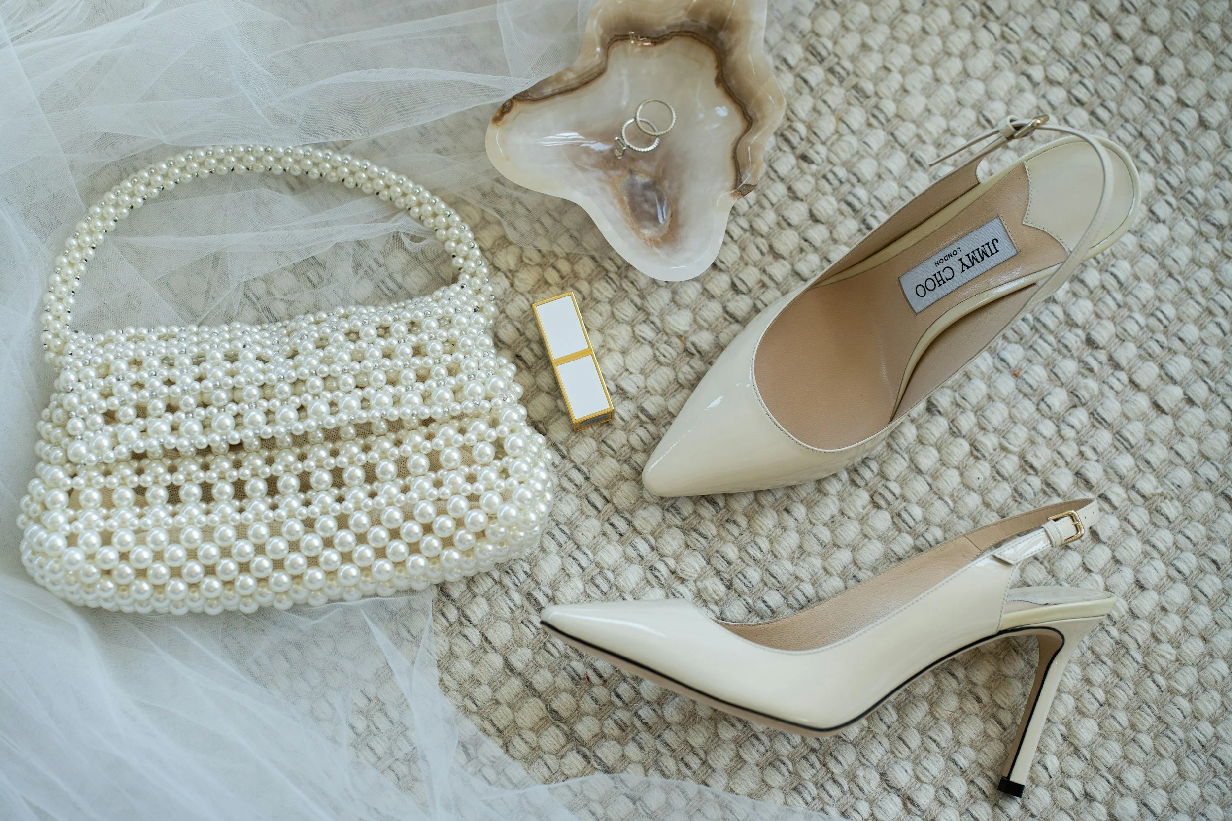 Elegante beige High Heels, Perlenschmuck, Ring, Make-up-Produkt, Perlentasche, und Dekorationen auf einer Teppichfläche