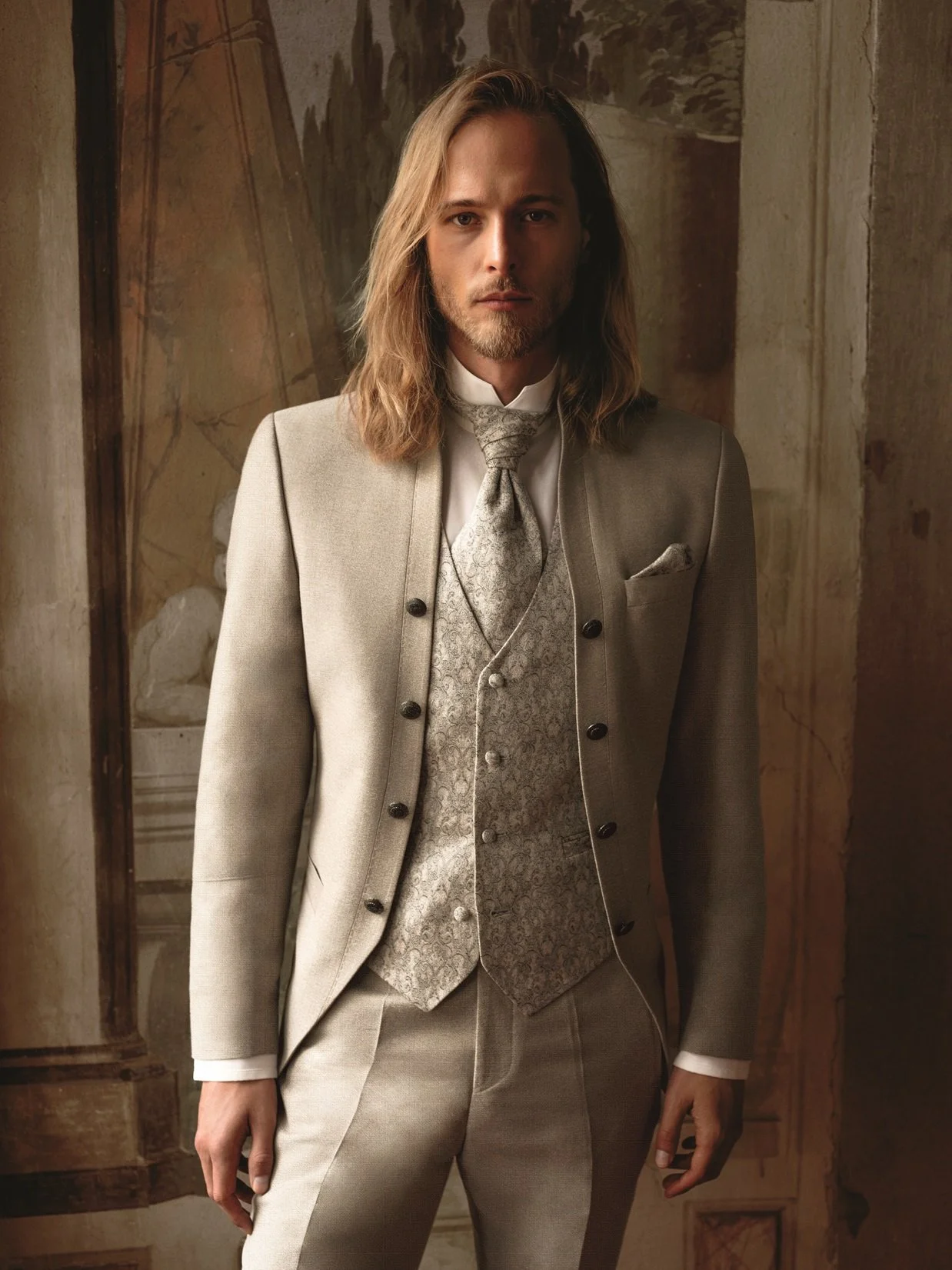 Ein Mann in einem eleganten, beige Anzug steht vor einer alten, restaurierten Wand mit Wandmalerei.