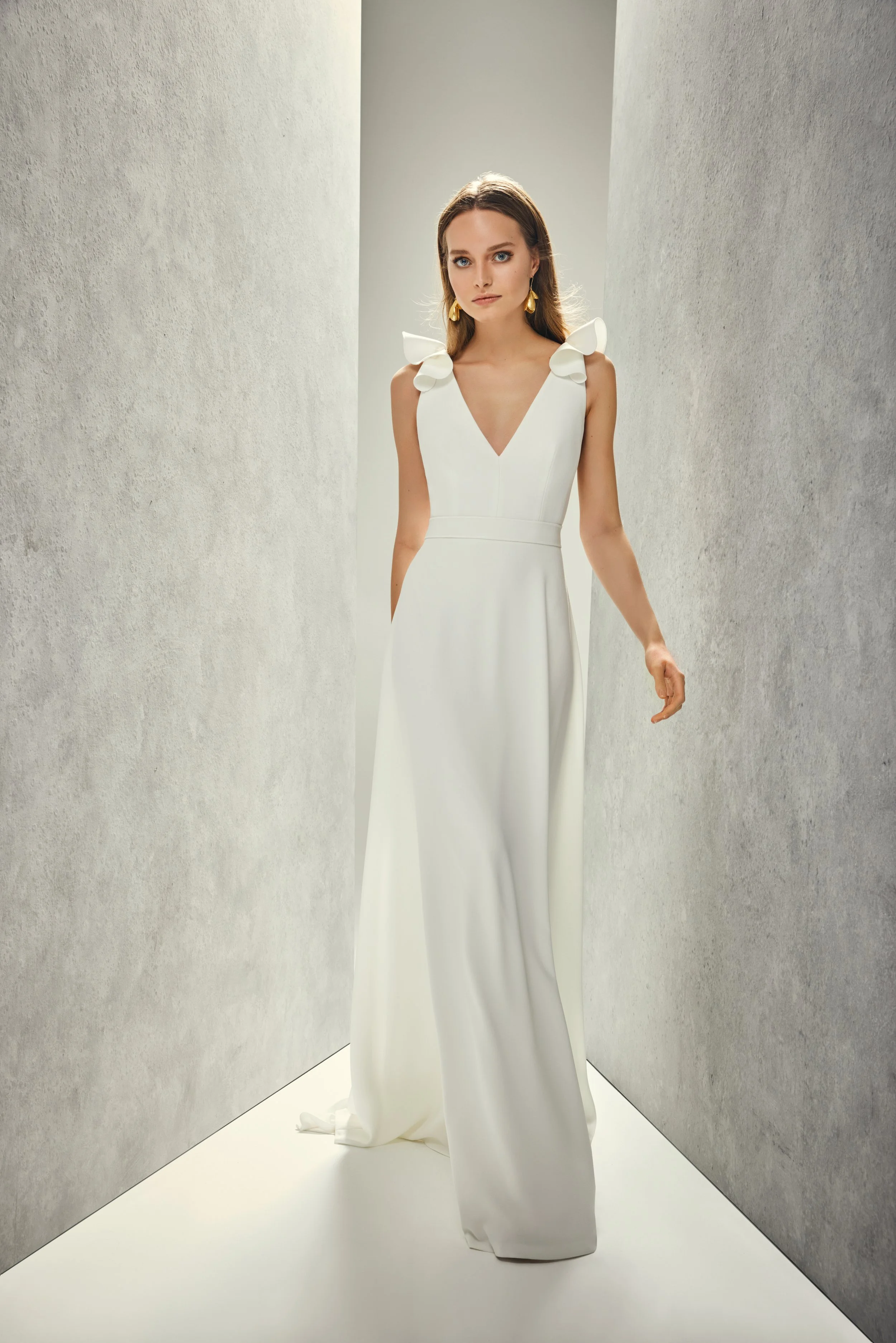 Ein Model in einem weißen Kleid mit Schulterdetails posiert in einem minimalistischen Raum mit grauen Wänden.