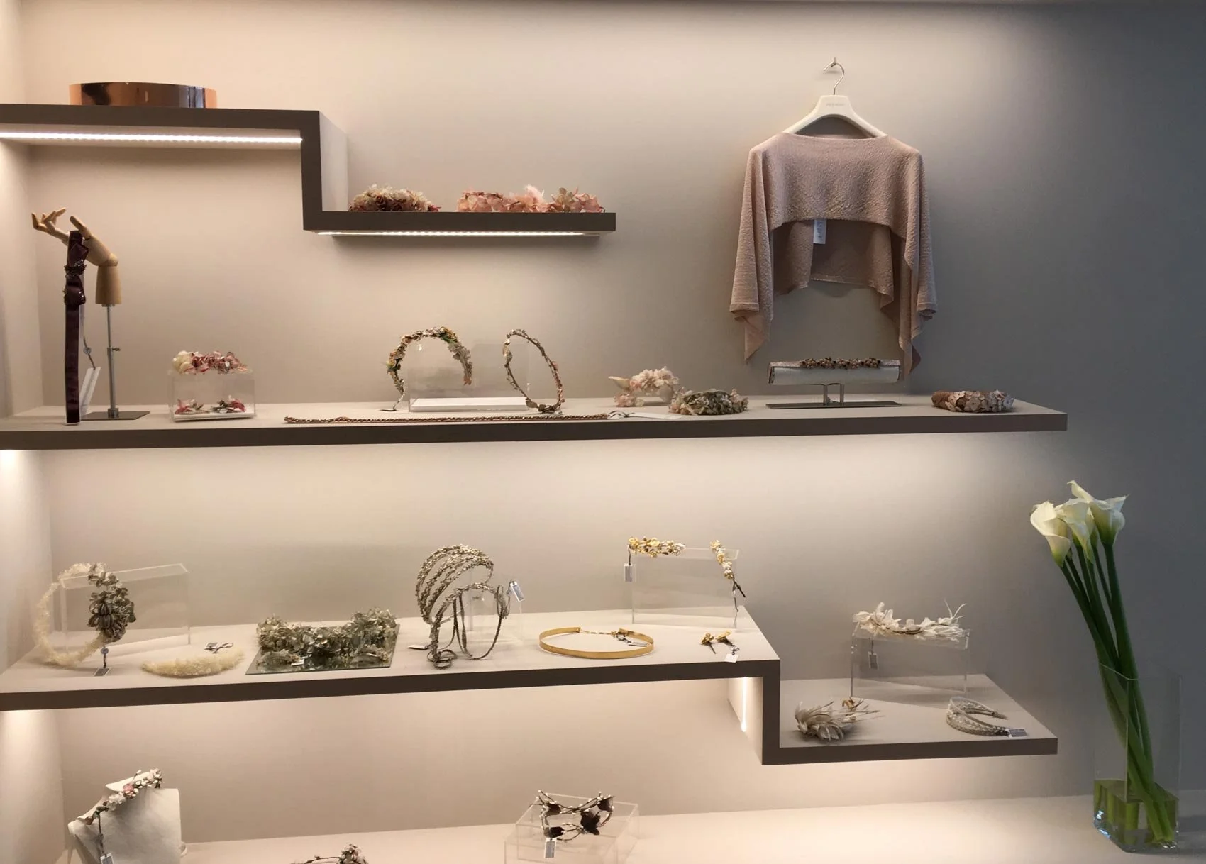 Ausstellung mit Schmuck und Accessoires auf mehreren weißen und grauen Ebenen, inklusive Kränze, Armbänder, Haarerker, und Kleidung auf einem Hängebügel, mit weißen Calla-Lilien in einer Vase auf der rechten Seite.