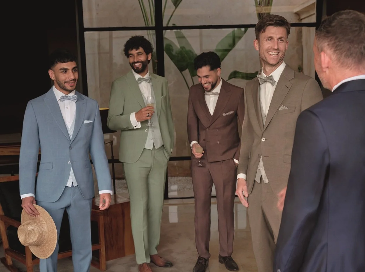 Fünf Männer in formeller Kleidung bei einer Party oder Veranstaltung, lachen und unterhalten sich in einem modernen, stilvollen Raum mit Pflanzen im Hintergrund.