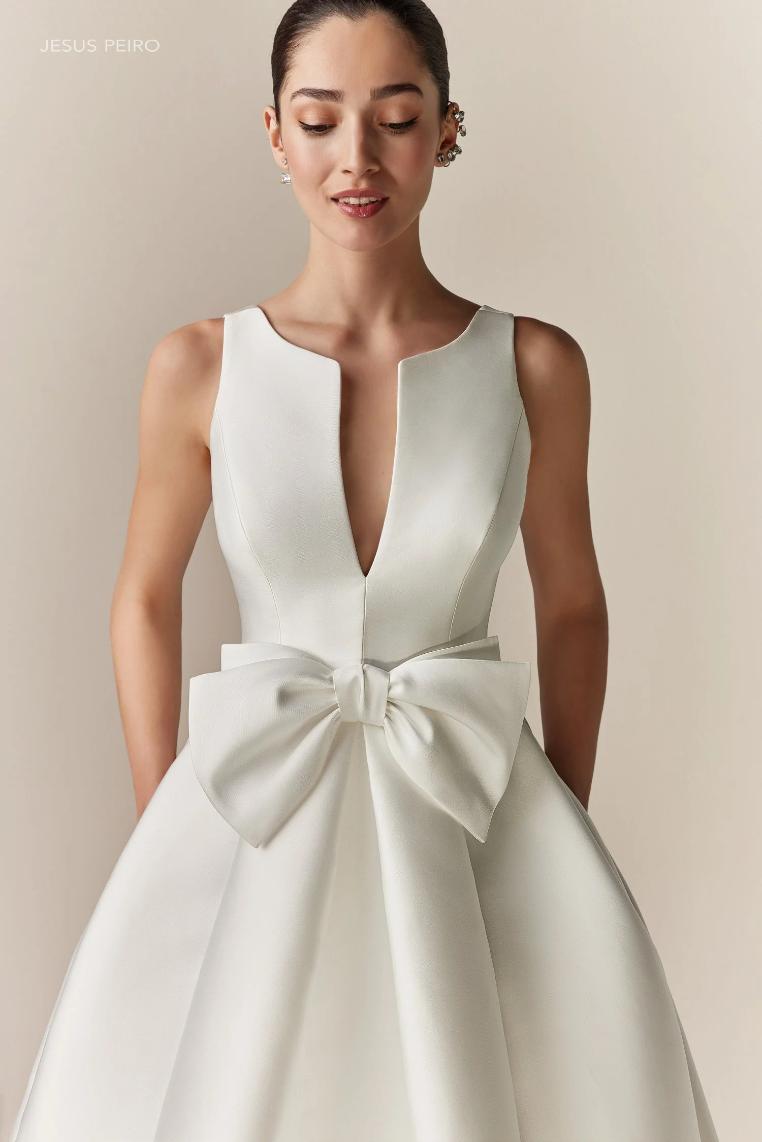 Ein junges Frau trägt ein elegantes, weißes Kleid mit einem großen, Stoffblumen-Detail an der Taille, mit glatten, dunkelbraunen Haaren und mehreren Ohrschmuck.