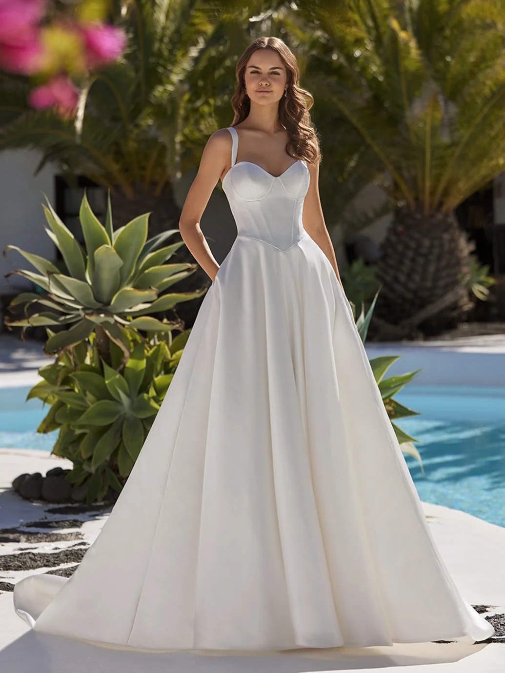 Frau in weißem Brautkleid vor Pool mit Palmen im Hintergrund.