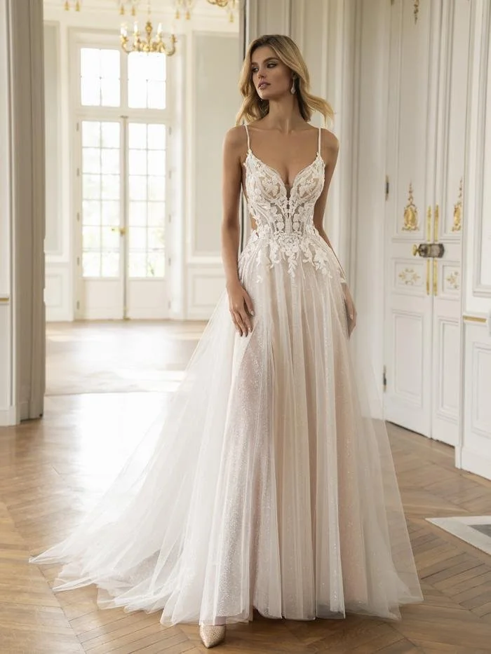 Eine Frau trägt ein elegantes, langes Hochzeitskleid mit Figur betonter Spitze und Tüll, in einem hell erleuchteten Raum mit weißen Wänden und großen Fenstern.