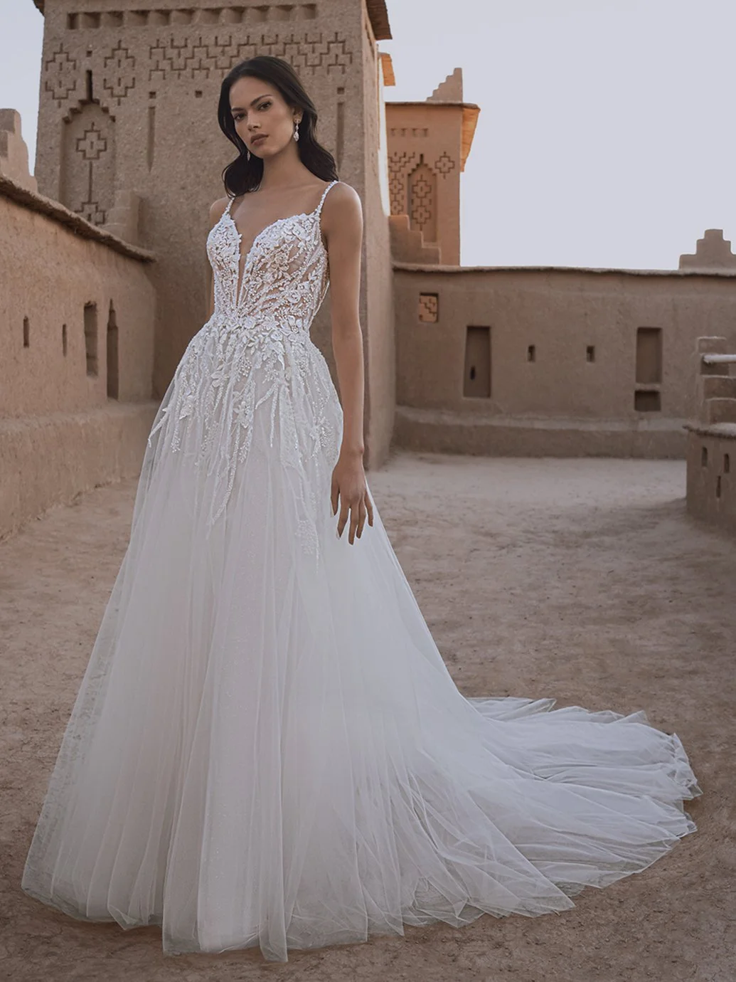 Frau in einem eleganten Hochzeitskleid mit Spitze und Tüll, vor einer alten, traditionellen Lehmburg im Wüstensand bei Sonnenuntergang.