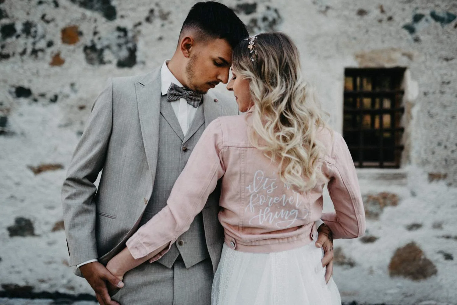 Ein verliebtes Paar steht eng nebeneinander vor einer alten Steinmauer, ihre Stirn aneinander, die Männer in einem grauen Anzug und die Frau in einem weißen Rock und einer rosa Jacke mit der Aufschrift "it's a forever thing".