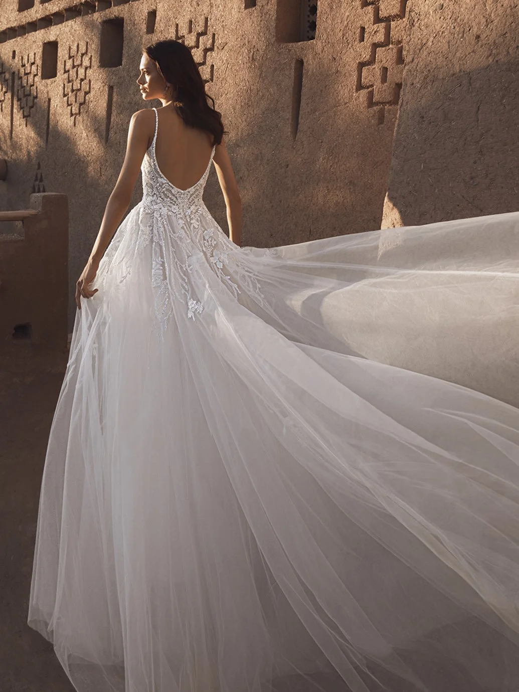 Eine Frau in einem weißen Brautkleid mit Spaghettiträgern und Tüllrock läuft vor einer roten Backsteinmauer. Sonnenlicht fällt auf sie.