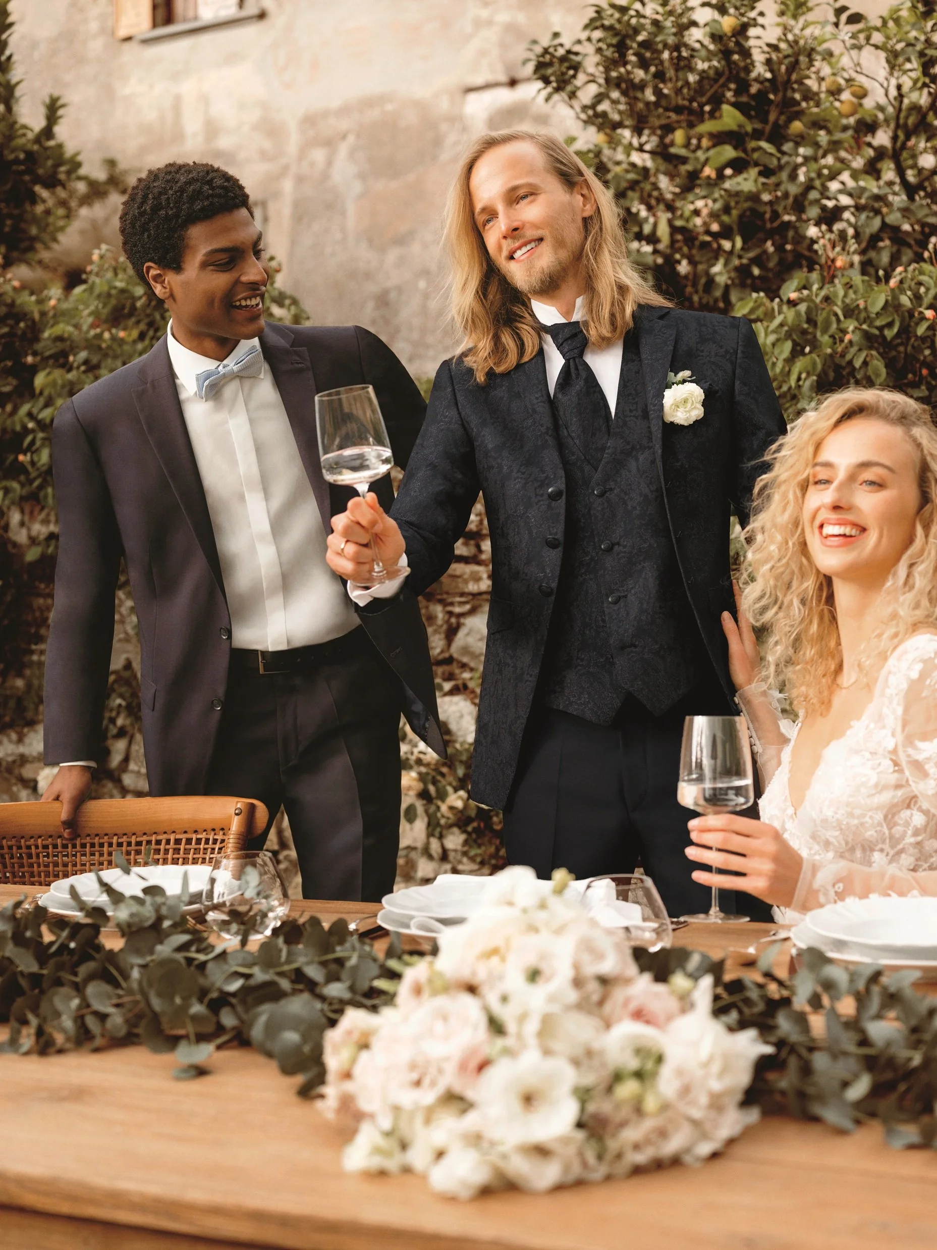 Menschen bei einer Hochzeitsparty, einer hält ein Weinglas, im Hintergrund eine Blumendekoration, sitzender Braut in Weiß, lachend, romantisches Ambiente