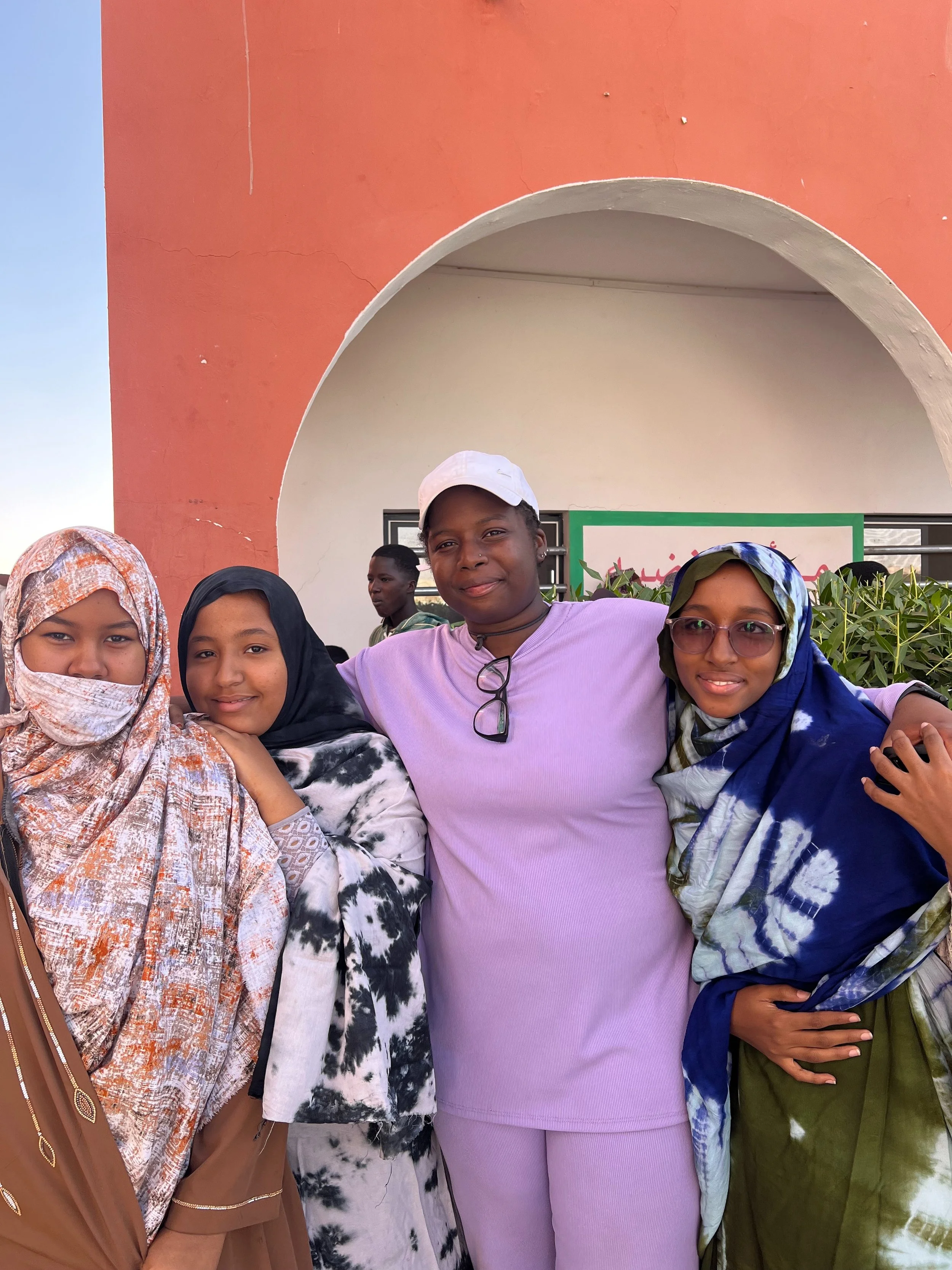 Groupe de femmes posant devant un bâtiment coloré, portant des vêtements traditionnels et modernes, avec un ciel clair en arrière-plan.