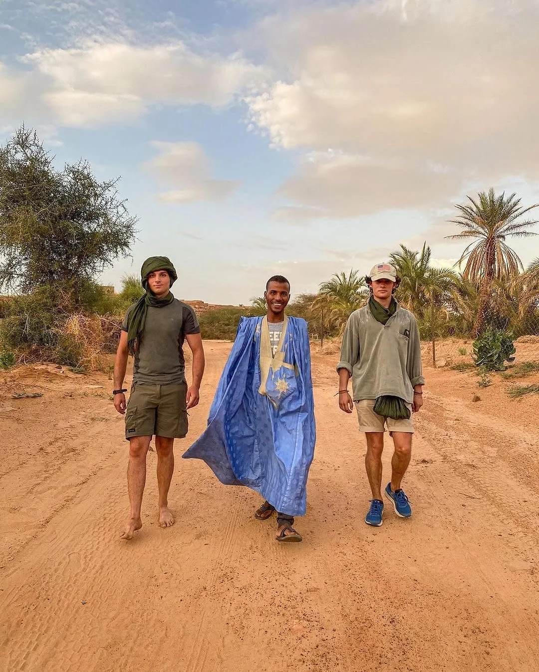 Trois jeunes hommes marchant dans un désert, l'un vêtu d'une robe bleue, avec des palmiers en arrière-plan et un ciel partiellement nuageux.