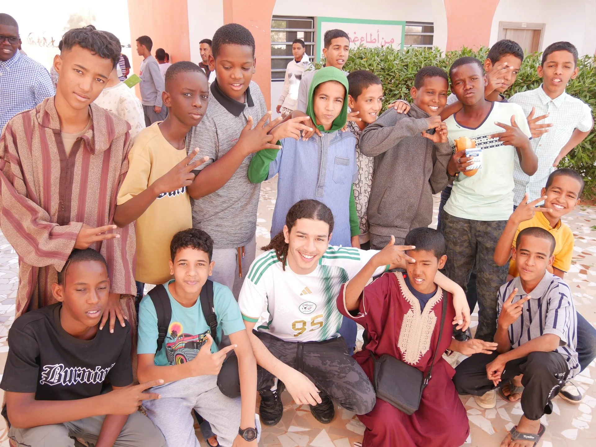 Groupe de jeunes garçons et filles posant pour une photo à l'extérieur, probablement devant une école, certains faisant des gestes avec leurs mains. Il y a des buissons en arrière-plan et un panneau en arabe dans la fenêtre.