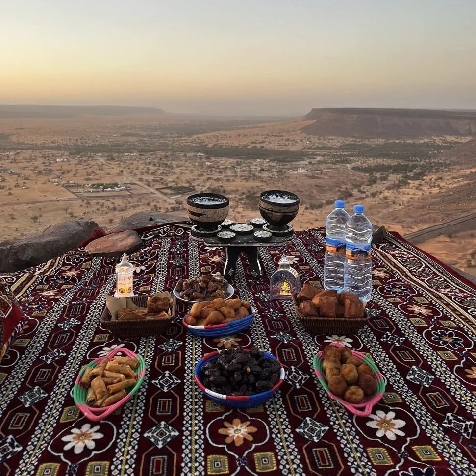 Repas traditionnel dans le désert avec vue panoramique sur les montagnes, comprenant des bols d'objets culturels, de la nourriture locale, des bouteilles d'eau et des décorations sur un tapis coloré.