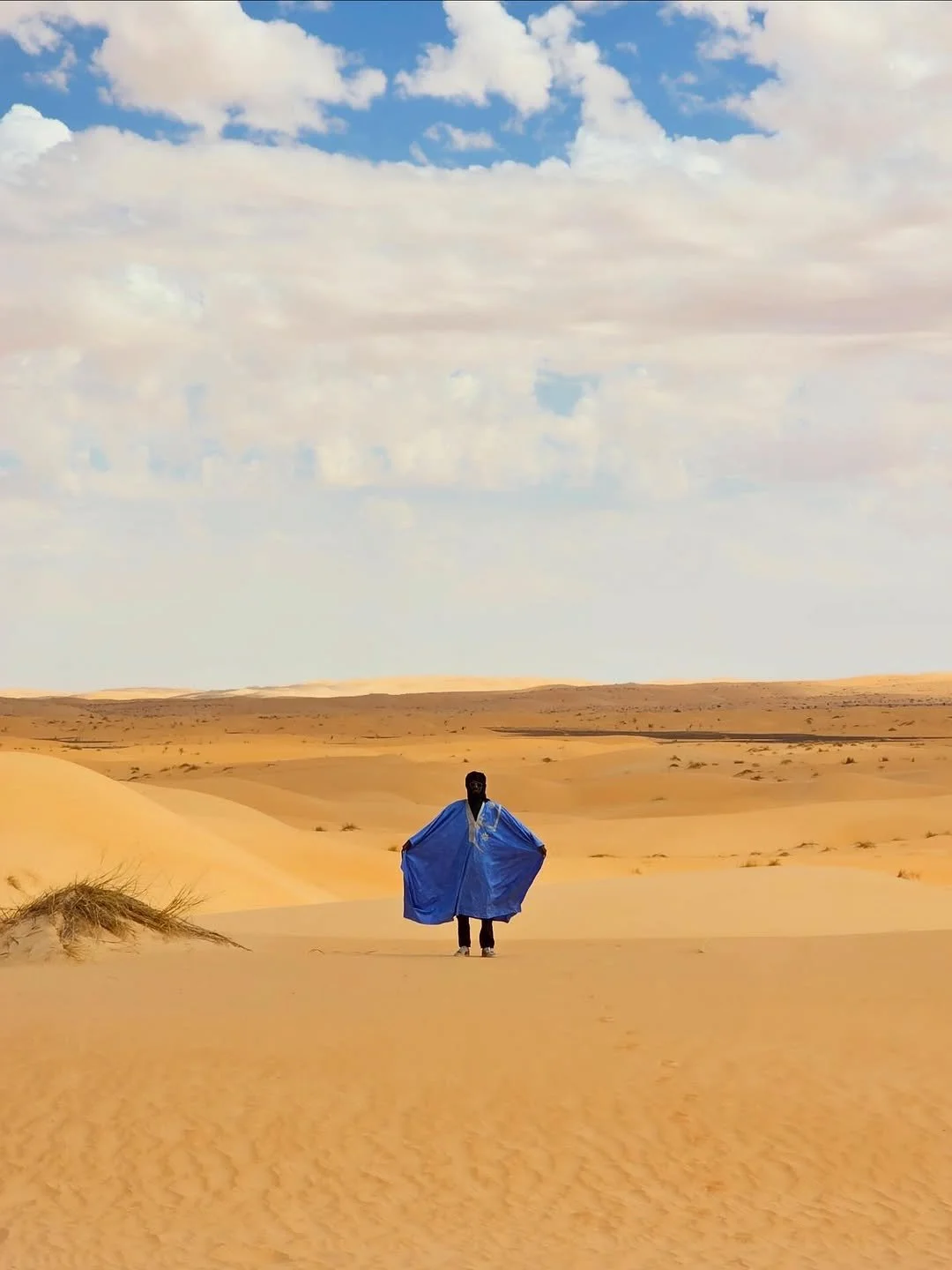Une personne portant une cape bleue dans un désert de dunes de sable avec un ciel partiellement nuageux.