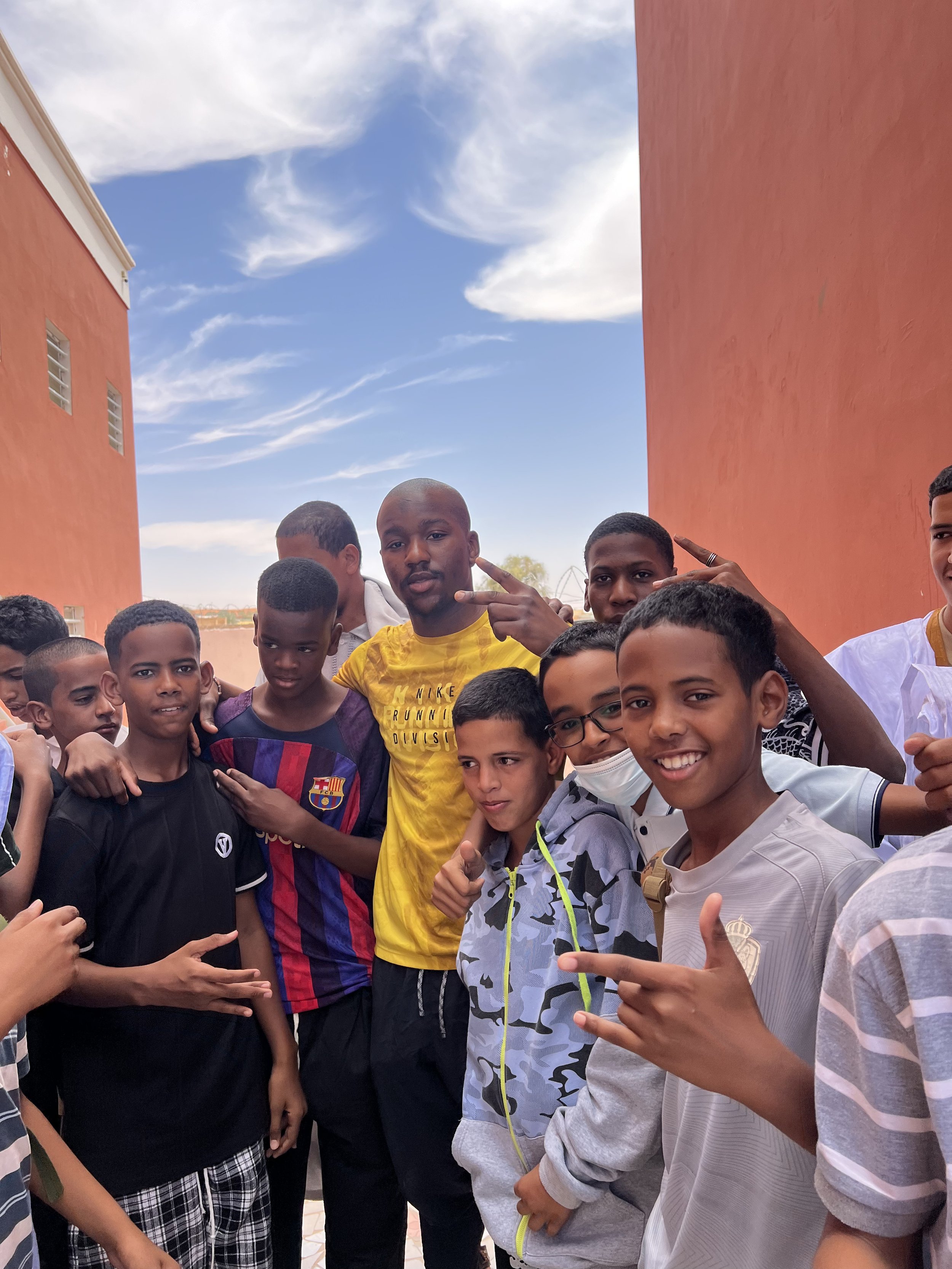 Groupe de jeunes garçons et jeunes hommes posant, certains faisant des signes de la paix, dans une cour extérieure sous un ciel bleu avec des nuages, entourés de bâtiments rouges.