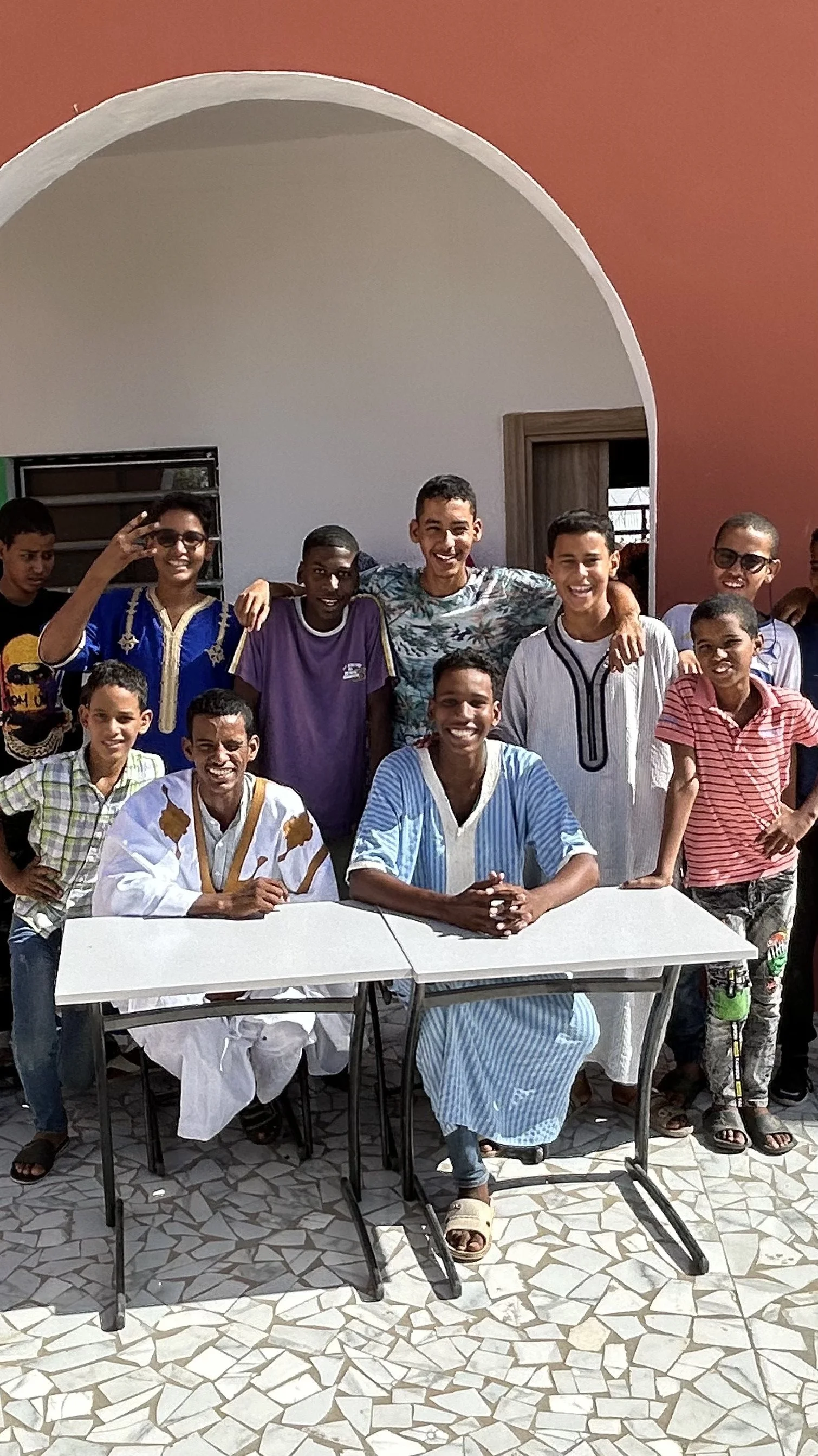 Groupe de jeunes et adultes africains posant pour une photo, certains souriants, dans une cour avec un sol en mosaïque. 