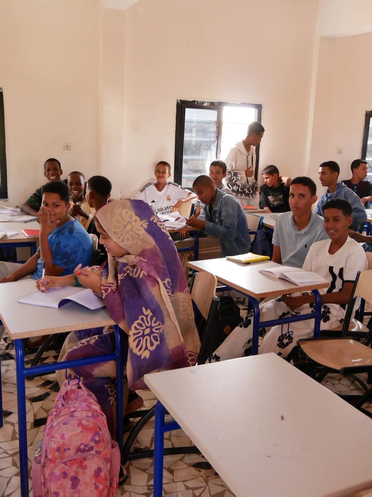 Des adolescents en classe, certains souriants, certains concentrés, avec des livres et des cahiers ouverts sur leurs bureaux, dans une salle lumineuse avec fenêtres.