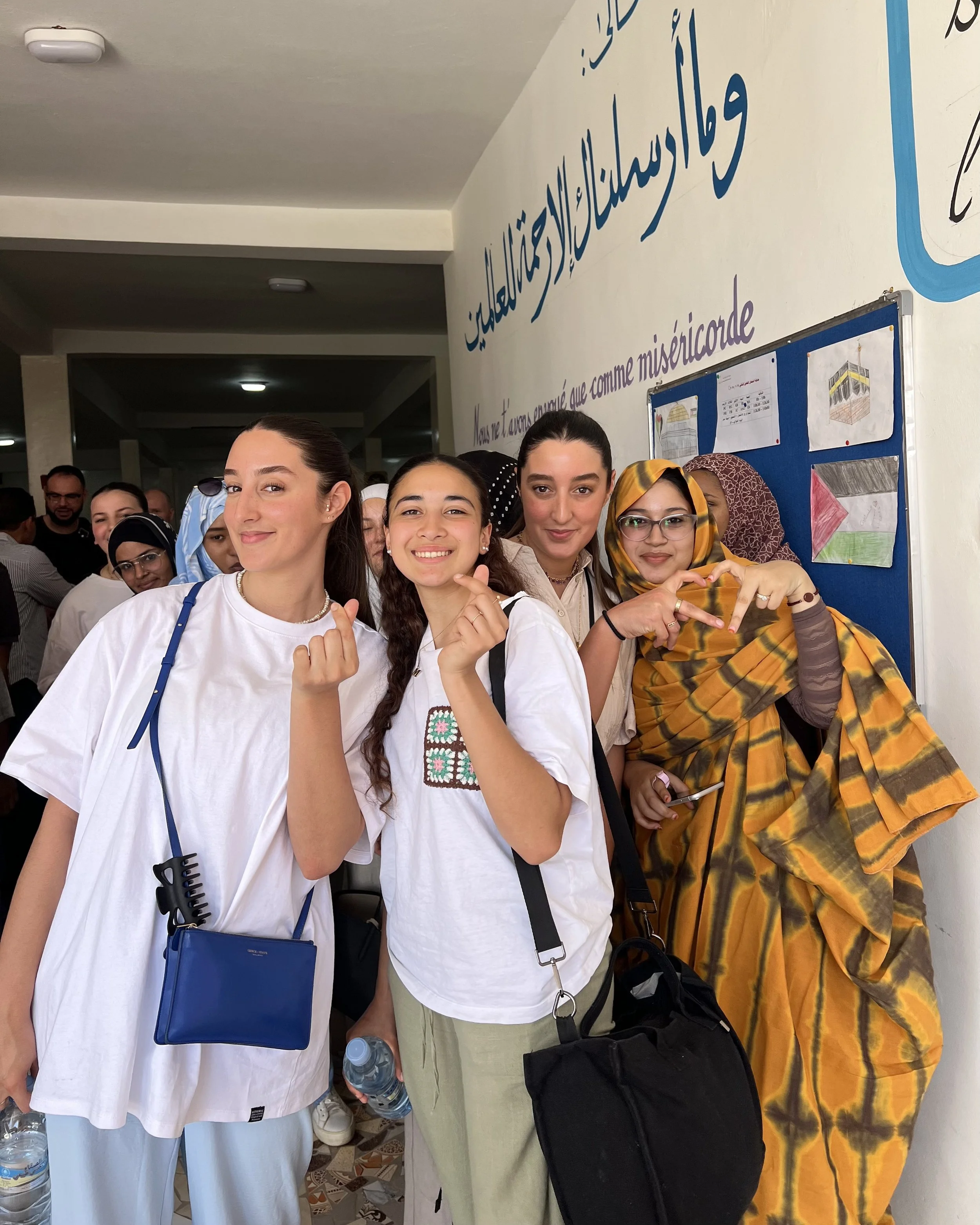 Groupe de jeunes femmes souriantes posant pour une photo lors d'un événement, avec un fond affichant du texte en arabe et des dessins colorés.