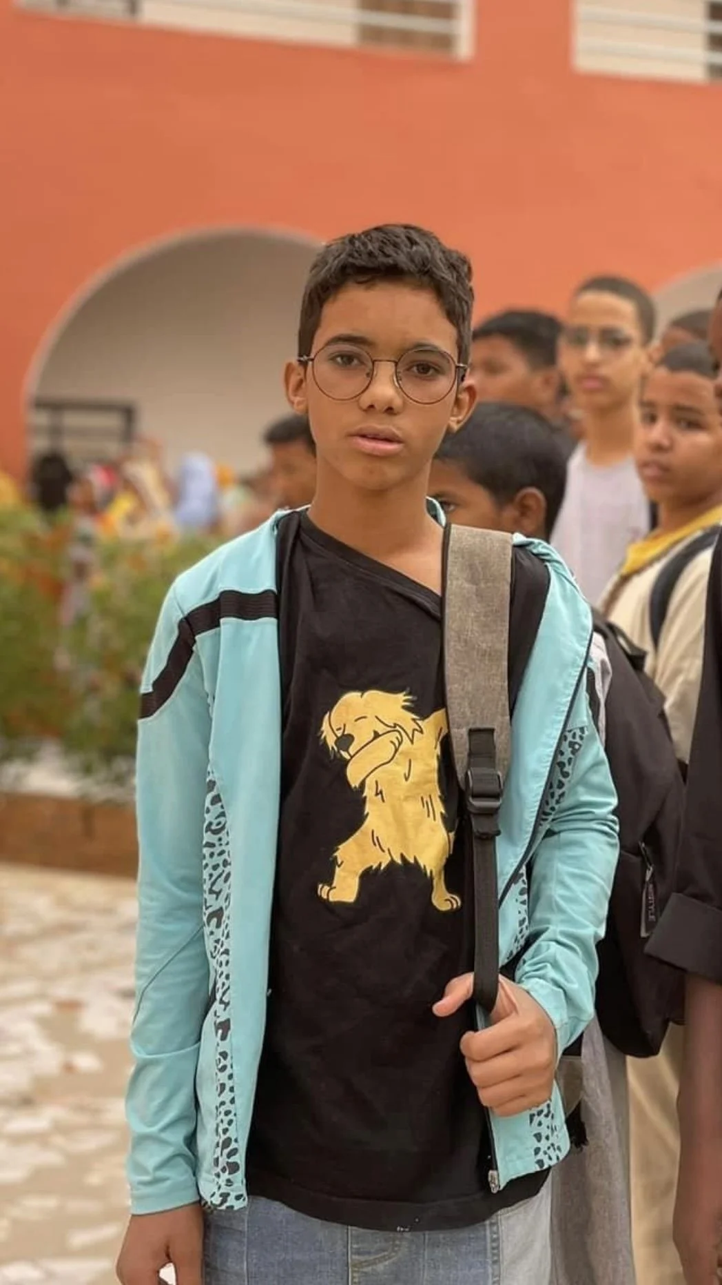 Un garçon portant une veste bleu clair et un t-shirt noir avec un lion en dessin, portant des lunettes, dans une foule devant un bâtiment orange.