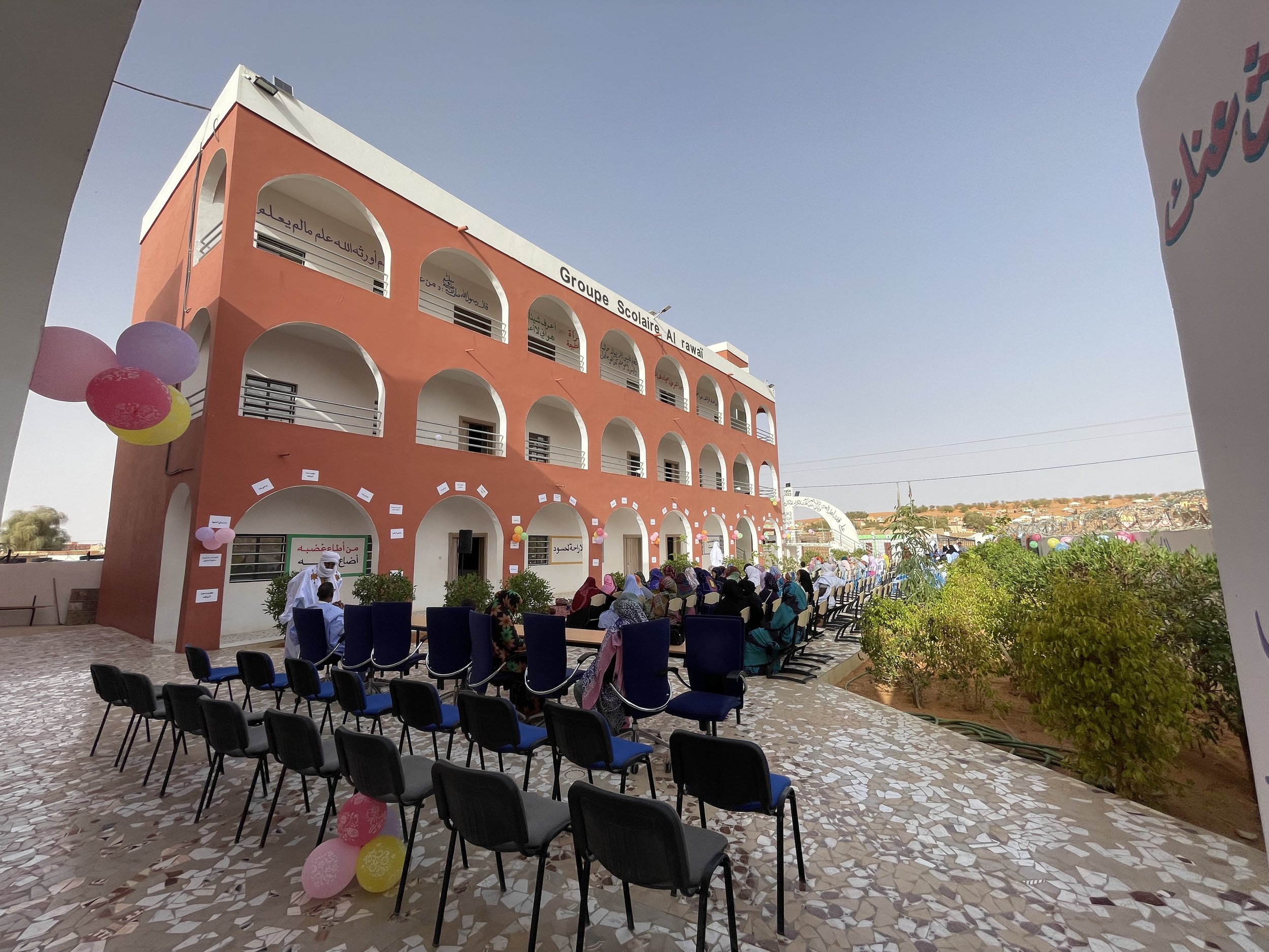 Une cour devant une école avec des chaises et des personnes assises, décorée avec des ballons colorés, bâtiment orange avec des arches et un ciel clair.