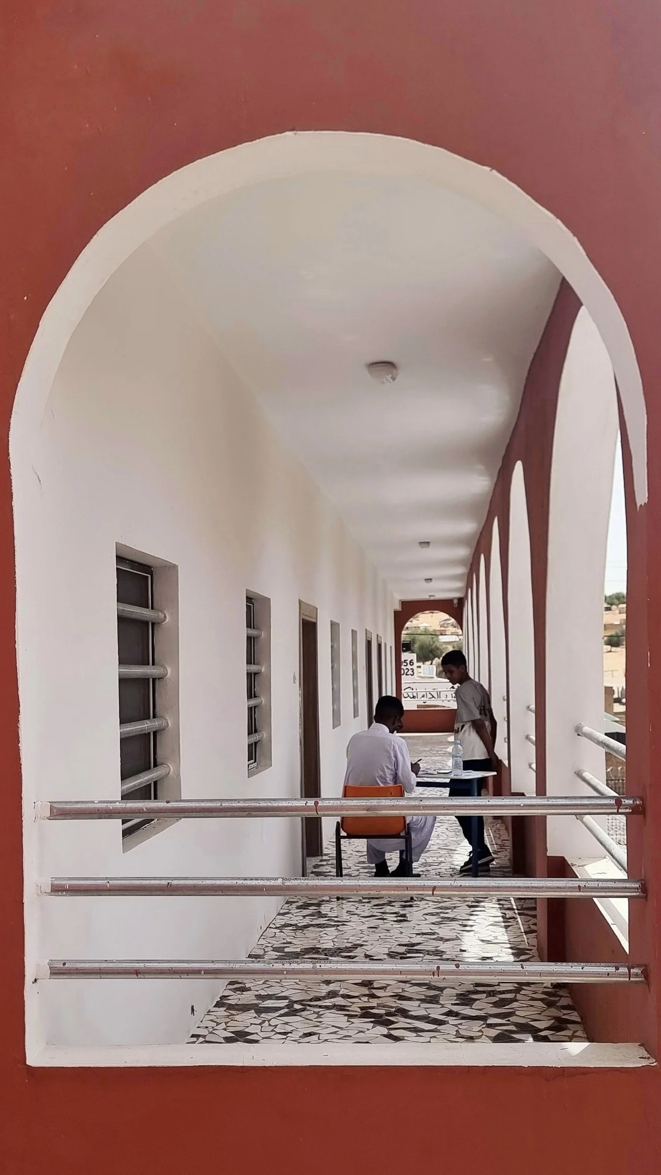 Une vue à travers une grille de sécurité montrant un couloir avec deux hommes, dont un assis et l'autre debout, discutant dans un bâtiment coloré avec murs blancs et arches rouges.