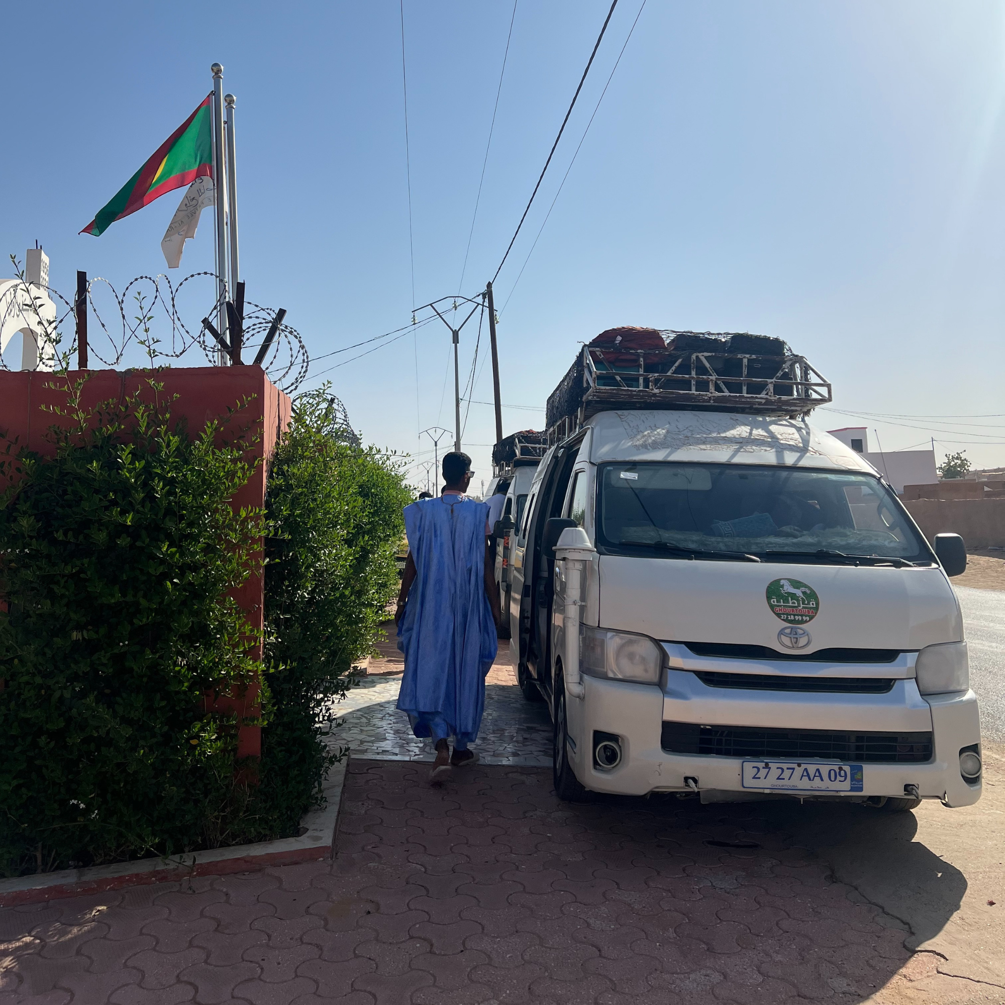 Un homme portant une robe bleue marche dans une rue où stationnent des voitures, avec des drapeaux flottant dans le ciel clair, un enroulement de fil de fer barbelé au sommet d'un mur à gauche, et un bâtiment avec un toit en béton noir.