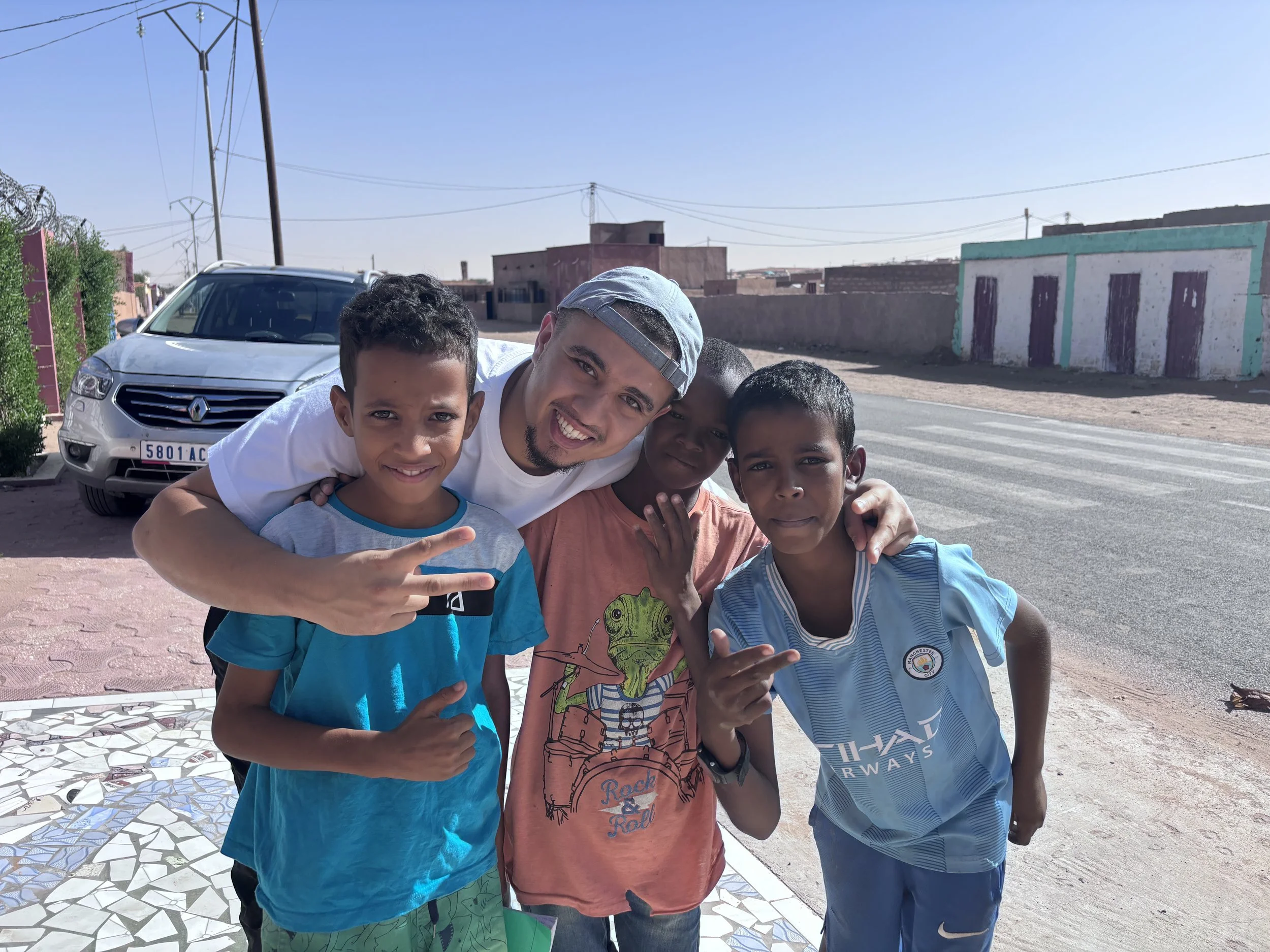Groupe d'un homme et trois enfants posant ensemble dans la rue en plein jour, avec des bâtiments en arrière-plan et une voiture blanche garée à côté.