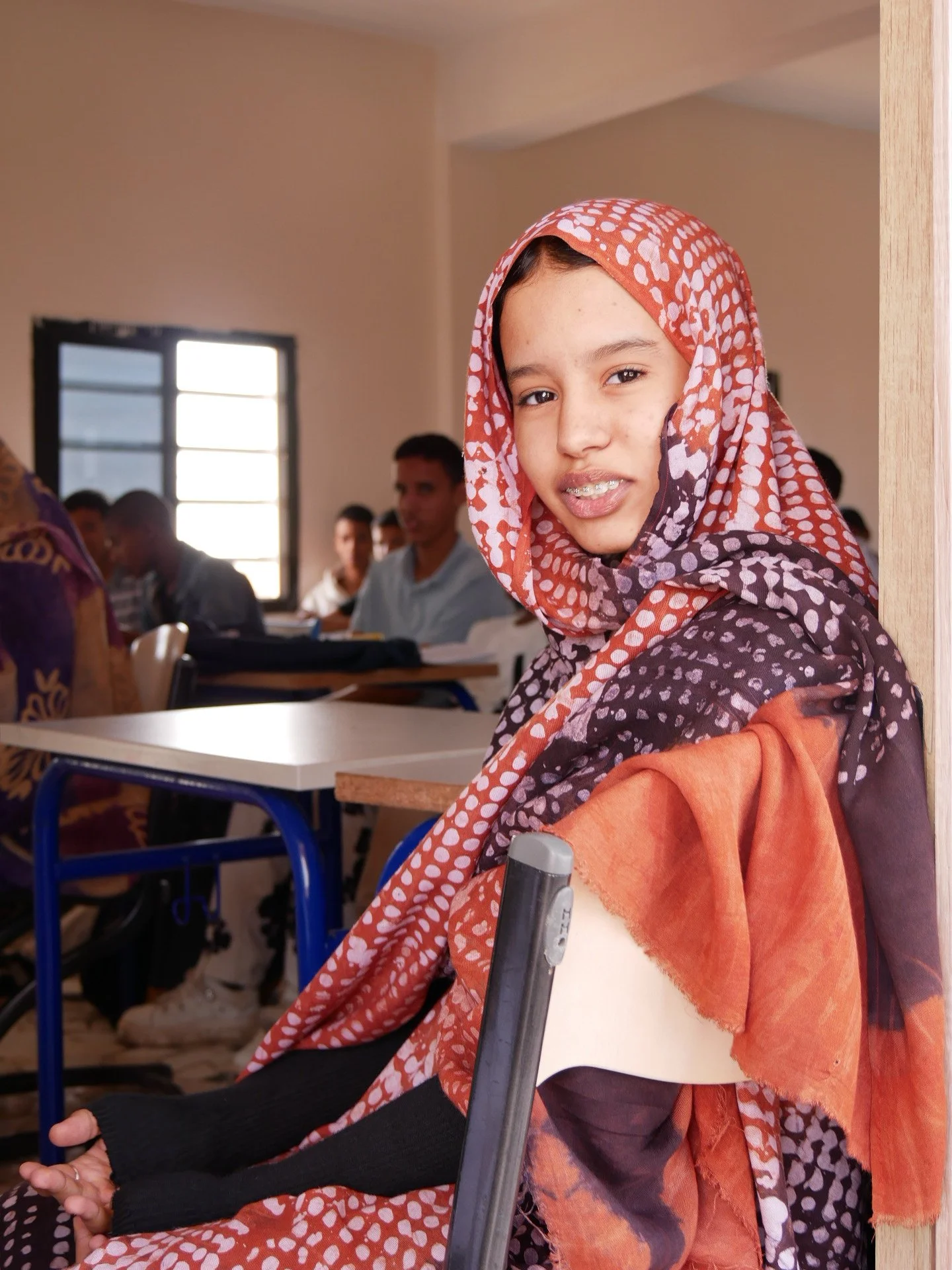 Une jeune fille porte un hijab à motifs rouges et blancs dans une salle de classe avec d'autres élèves en arrière-plan.
