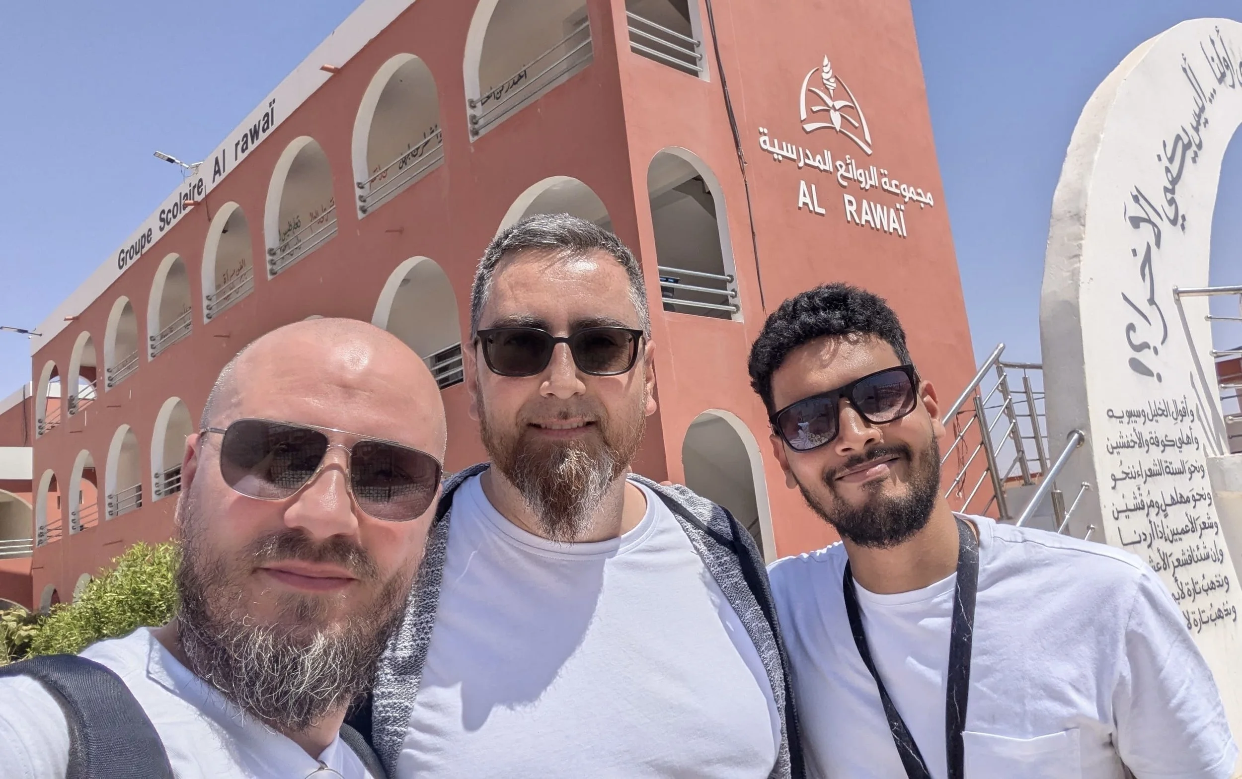 Groupe de trois hommes portant des lunettes de soleil devant un bâtiment appelé 'Al Rawā’'.