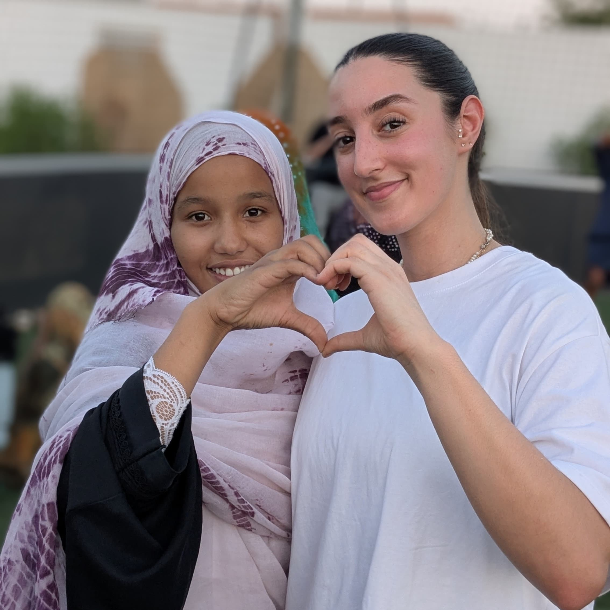 Deux personnes, une enfant avec un foulard rose et une femme avec des boucles d'oreilles, forment un cœur avec leurs mains.