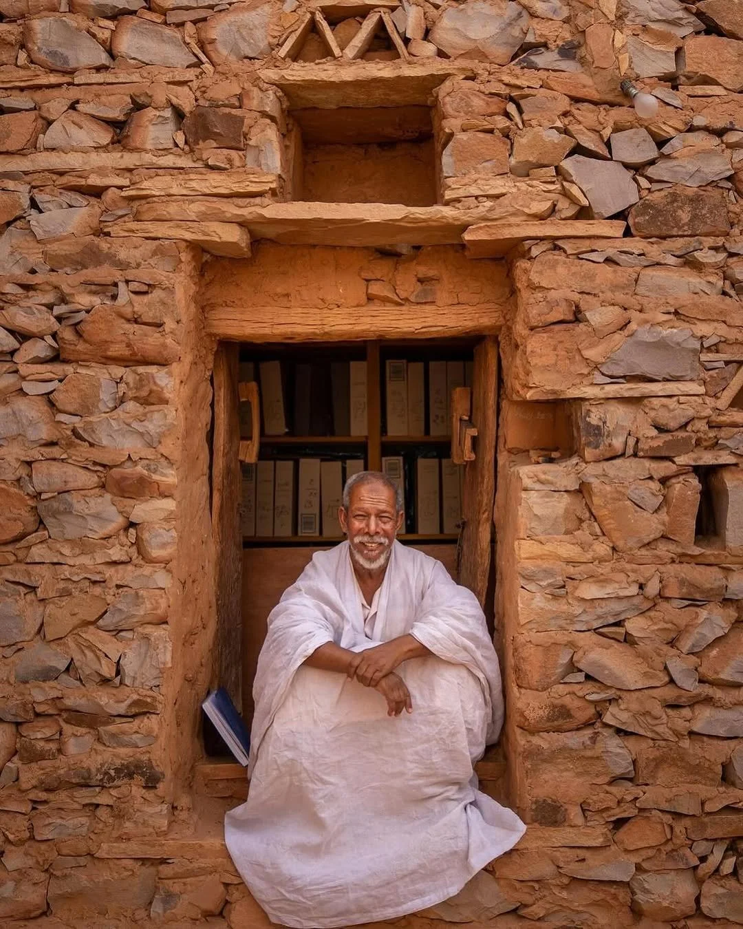 Un homme âgé souriant, assis dans une fenêtre encadrée de pierre, portant une robe blanche, avec un fond de rayonnage de livres dans un mur en pierre.