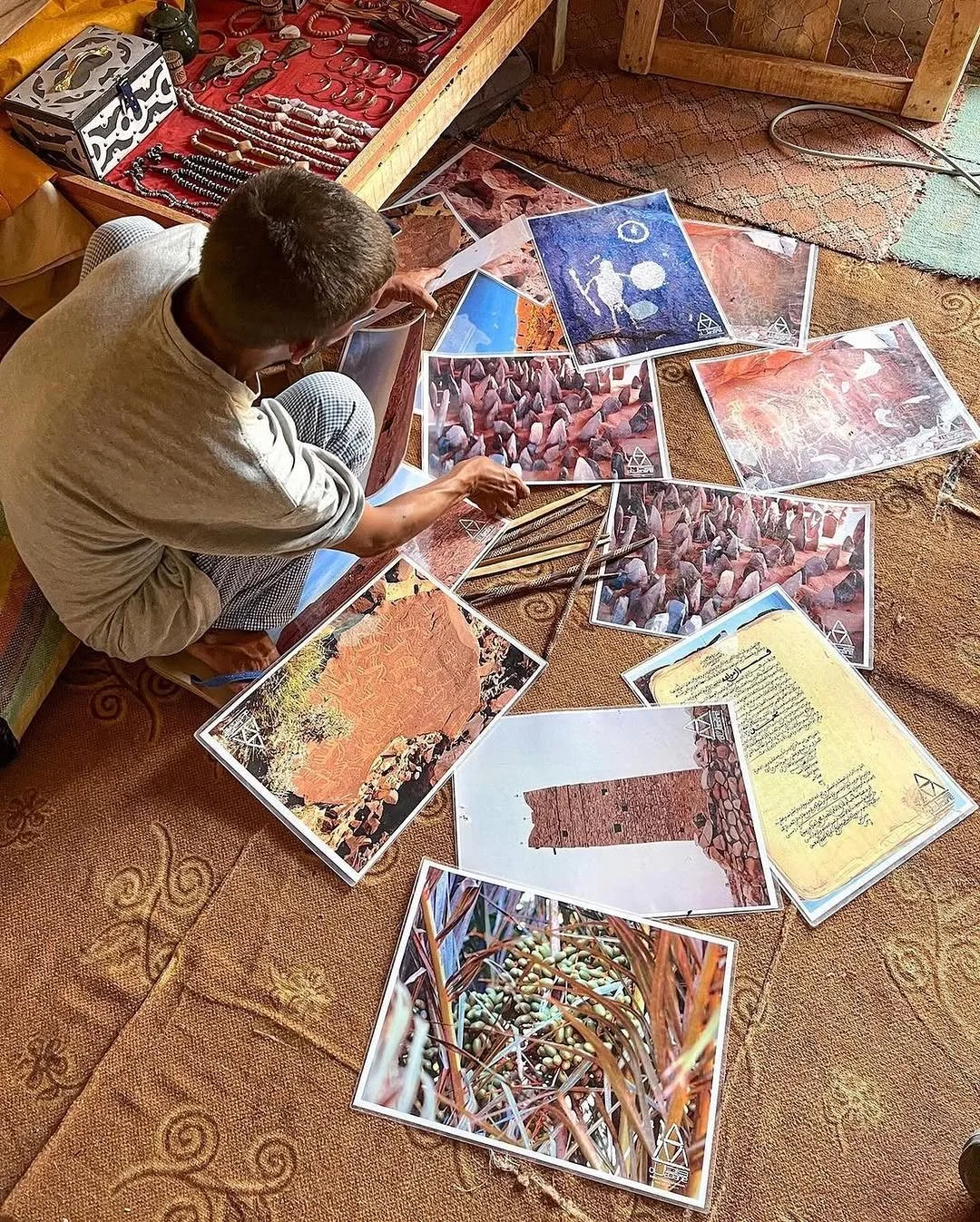 Une personne assise sur un tapis en train de sortir des photographies colorées et une carte, entourée de plusieurs autres photos au sol, avec une étagère à bijoux en arrière-plan.