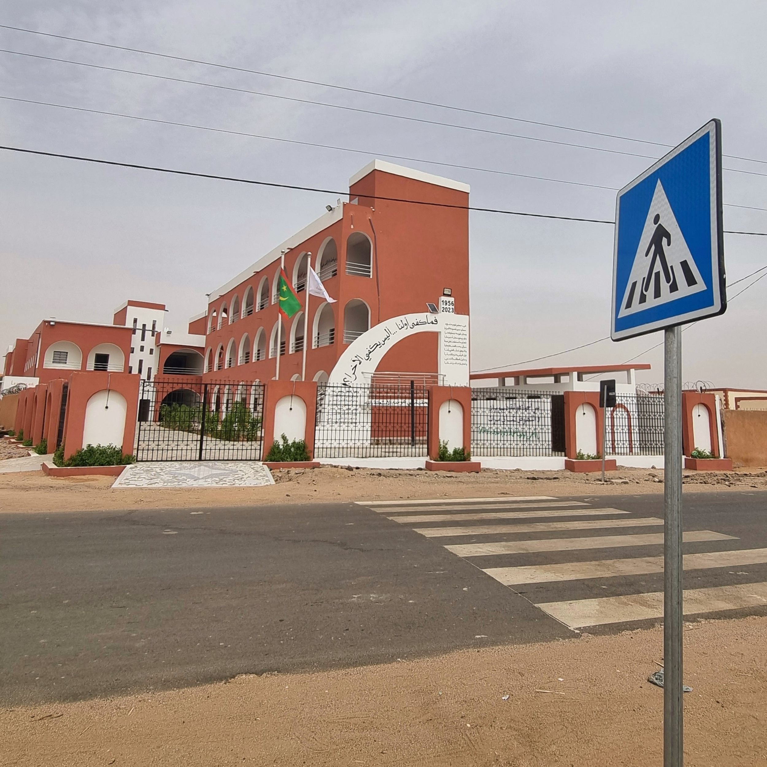Une rue avec un passage piéton devant un bâtiment coloré en orange avec plusieurs drapeaux, un panneau de signalisation pour passage piéton, et un ciel nuageux.