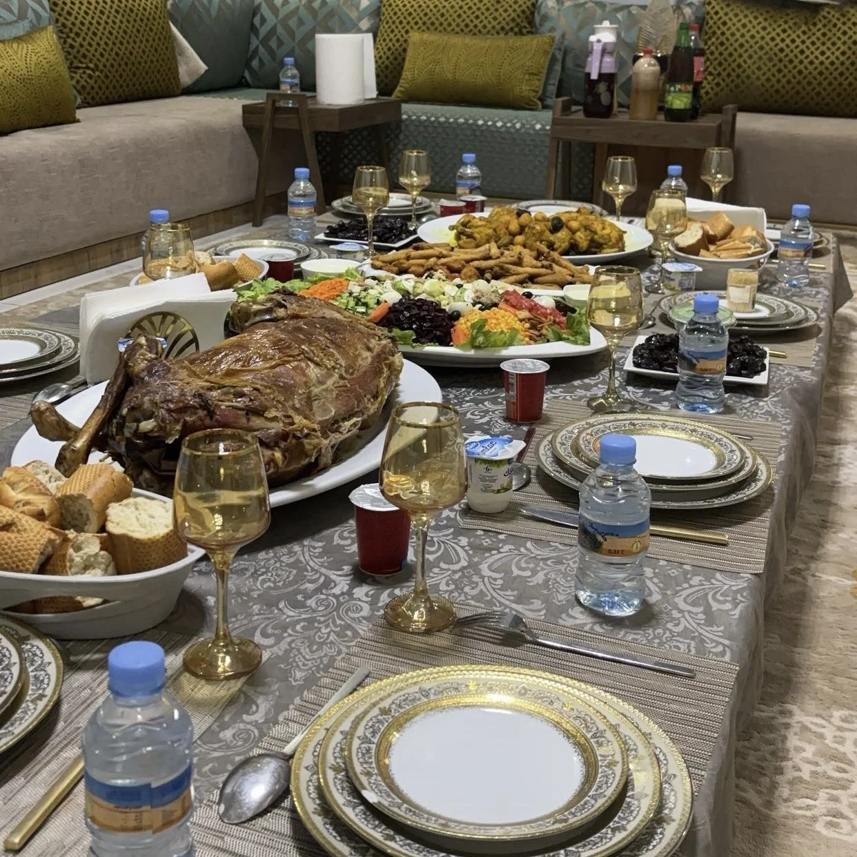 Table dressée avec un grand festin comprenant une pièce de viande, des salades, des brochettes, des plats de légumes, des boissons, des pâtisseries et des bouteilles d'eau, dans une salle décorée avec des coussins sur un canapé.