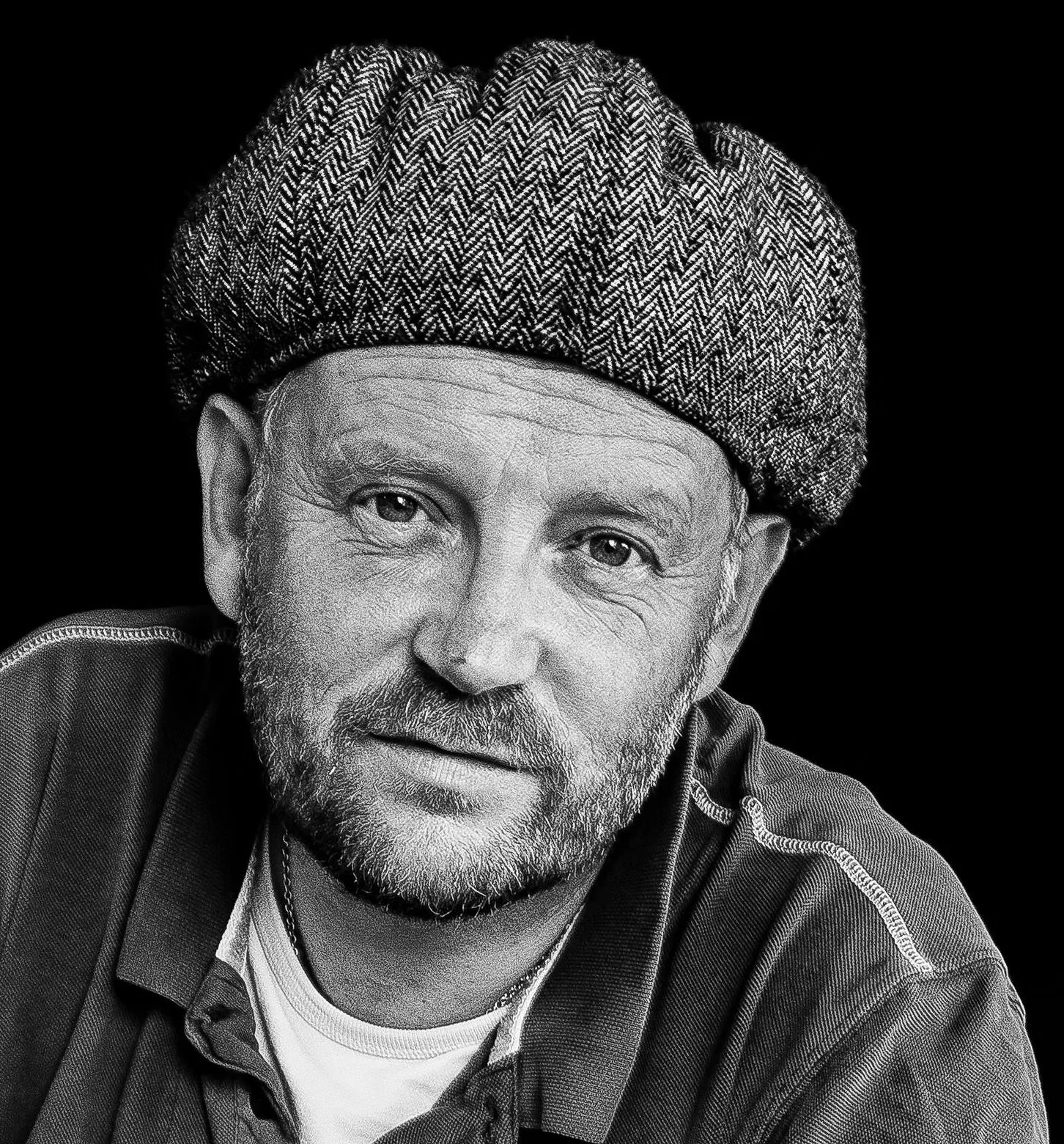 Black and white close-up portrait of an older man with a beard, wearing a patterned beret and a casual shirt, against a plain dark background.