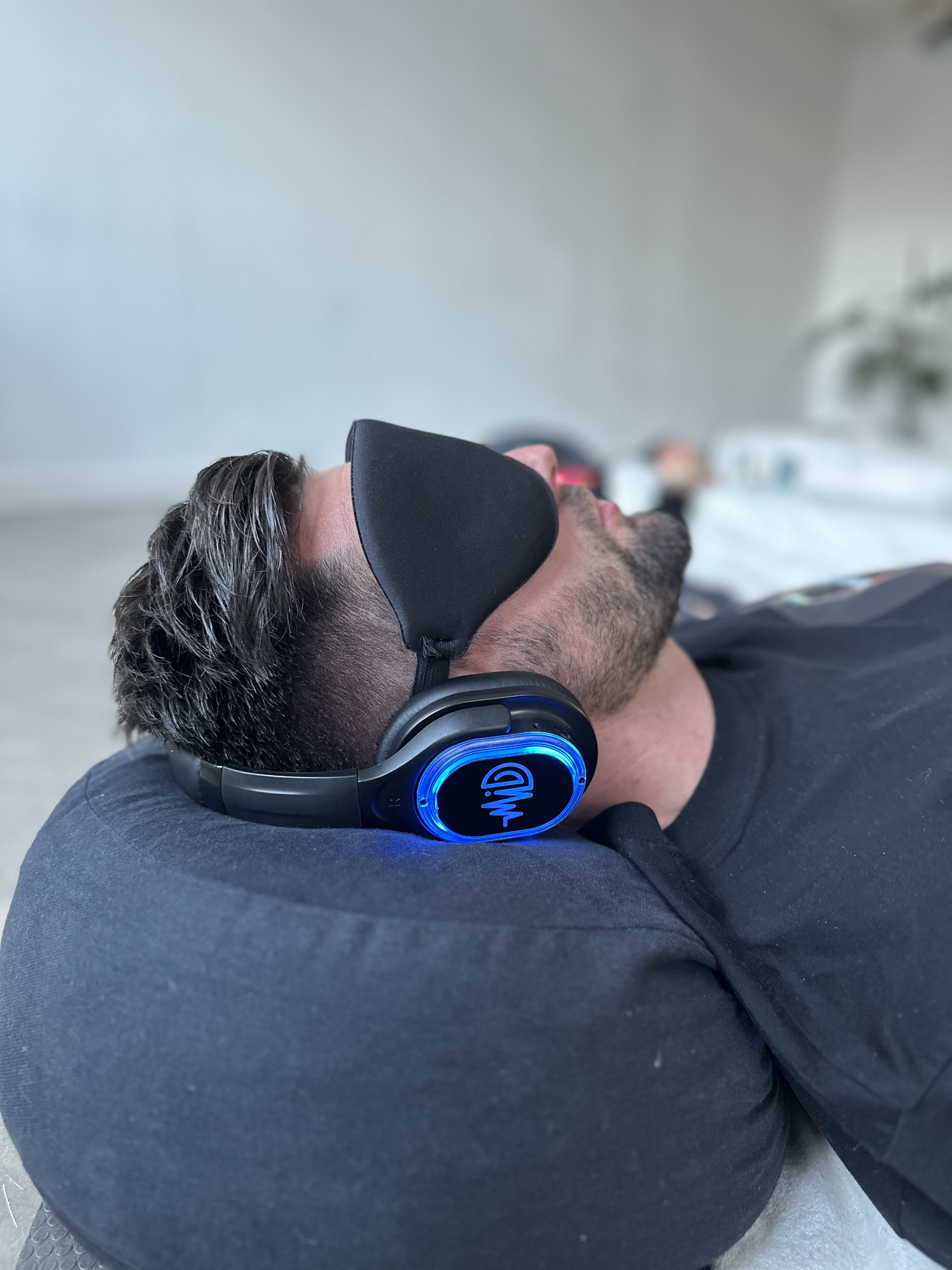 A man with short brown hair and a beard wearing black headphones and a black sleep mask relaxing on a grey surface.