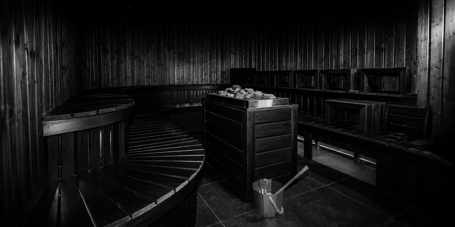 Black and white photo of an empty sauna room with wooden benches and a wood stove in the center.