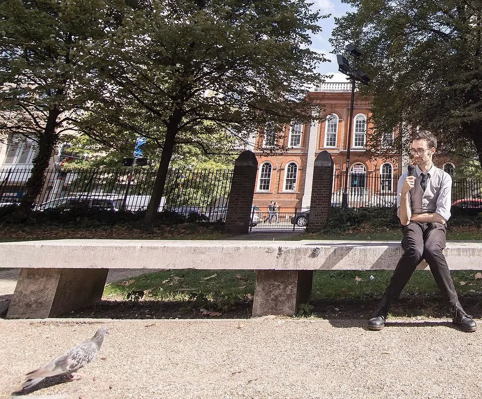 A person in professional attire, sitting on a concrete park bench and holding a microphone, is outdoors in front of a historic brick building with trees and a fence surrounding the area. A pigeon is on the ground nearby.