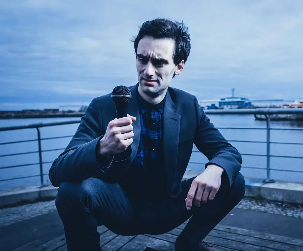 Man in a suit crouching outdoors holding a microphone with a body of water and cloudy sky in the background.