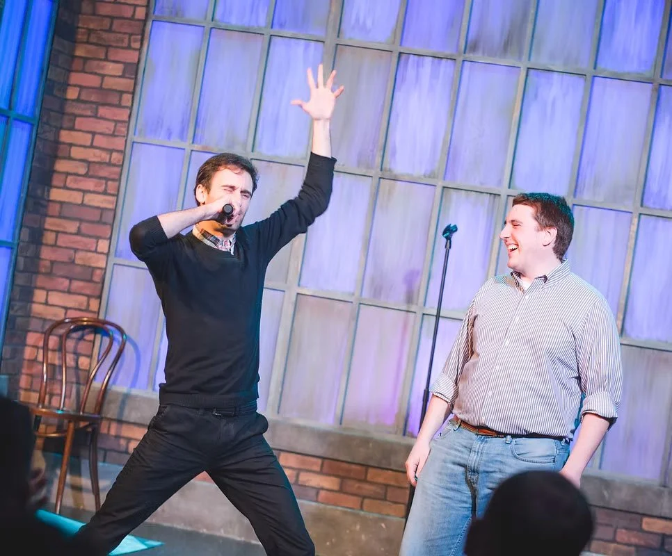 Two men on stage, one singing into a microphone with one arm raised, the other smiling and looking at him, in a setting with brick wall and large window panels.