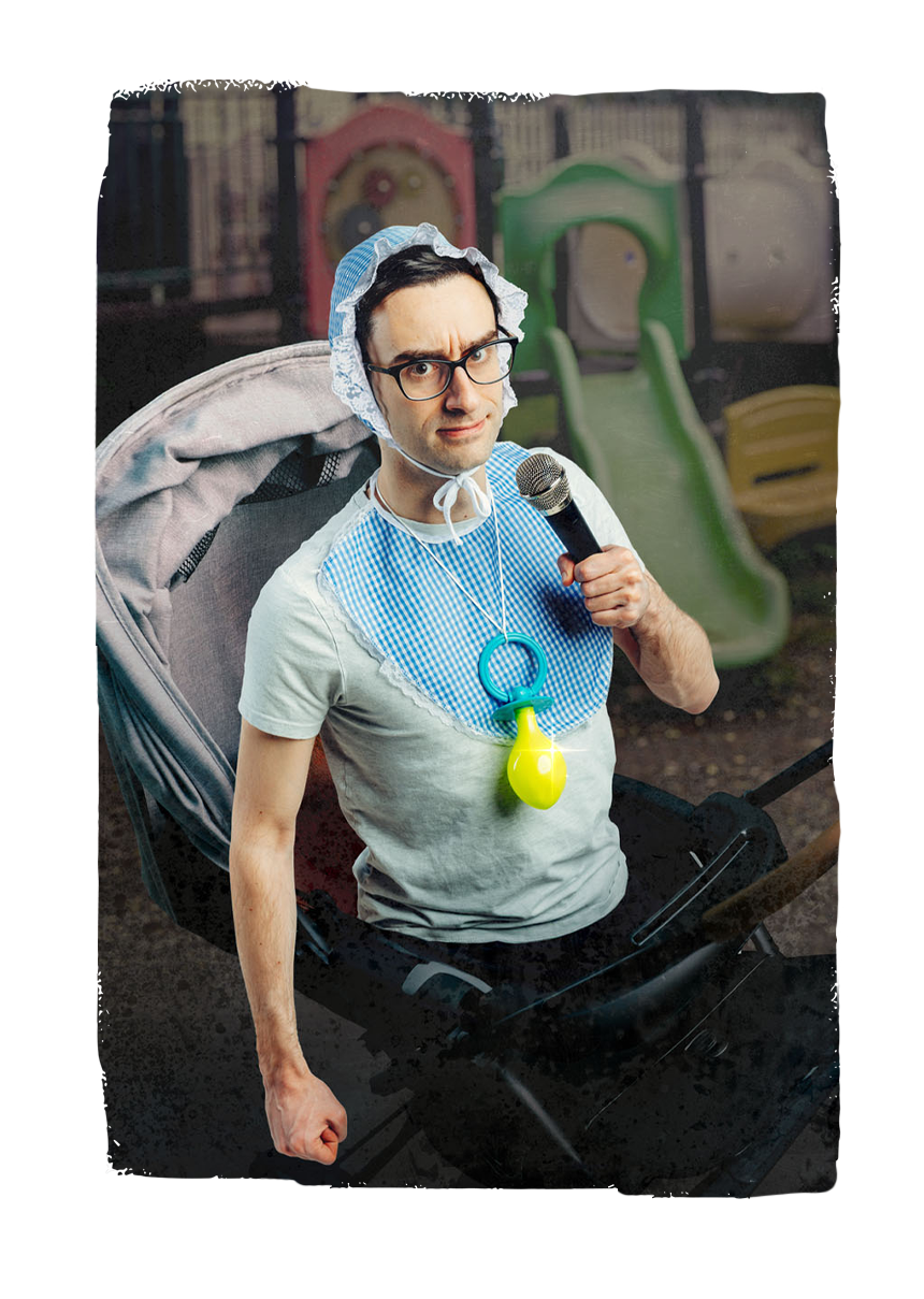 A man dressed as a baby, with a pacifier, baby bib, and bonnet, holding a microphone and carrying a stroller, standing in front of a playground.